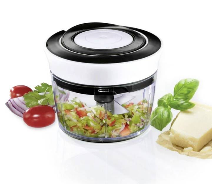 A vegetable chopper with chopped vegetables inside, surrounded by cherry tomatoes, onions, herbs and a piece of Parmesan cheese on paper.