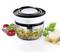 A vegetable chopper with chopped vegetables inside, surrounded by cherry tomatoes, onions, herbs and a piece of Parmesan cheese on paper.