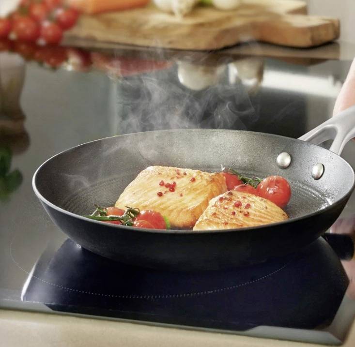 Two salmon fillets and cherry tomatoes are sizzling in a pan on the hob. Steam rises, with a chopping board visible in the background.