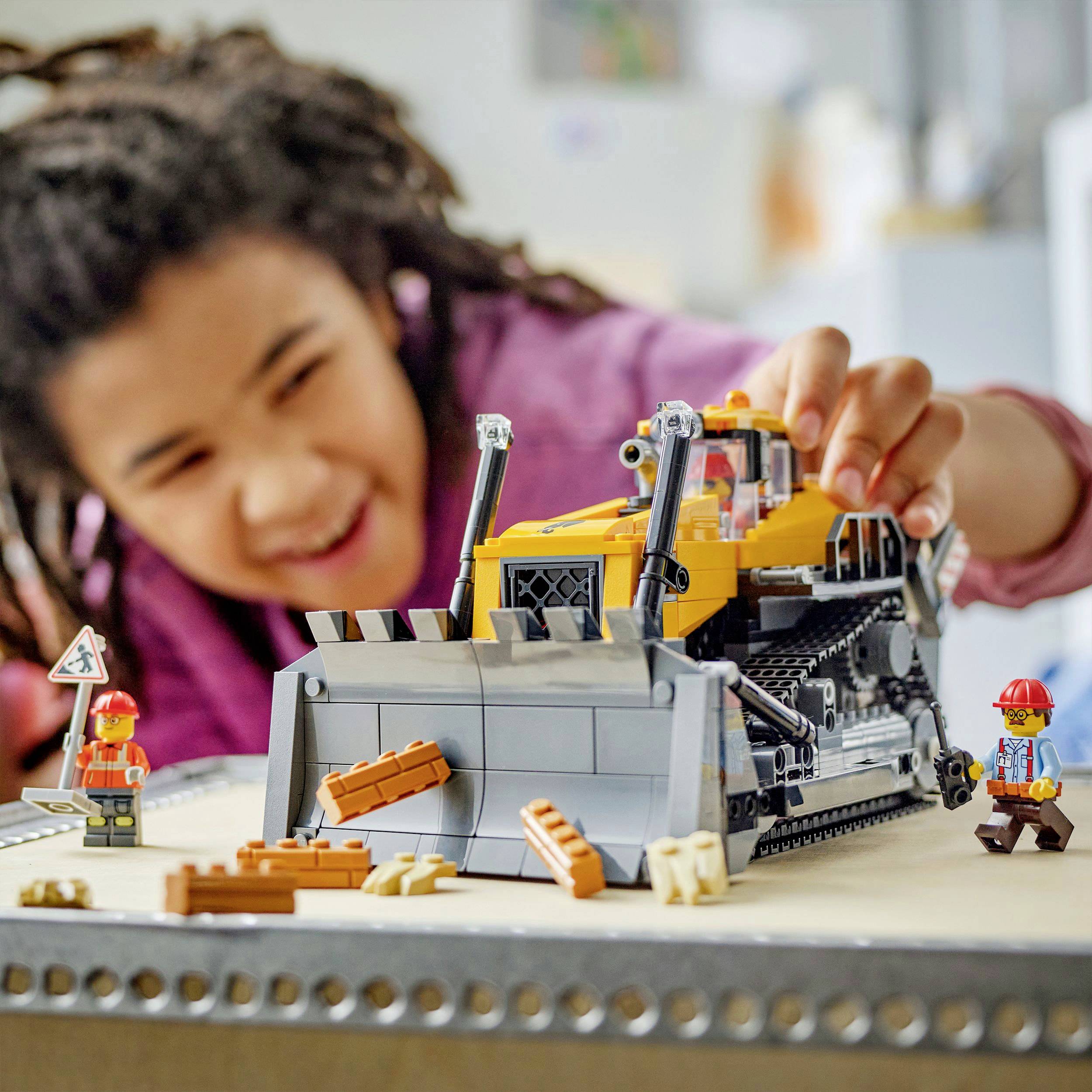 A child plays happily with a toy bulldozer on a table, surrounded by small toy figures and plastic parts.