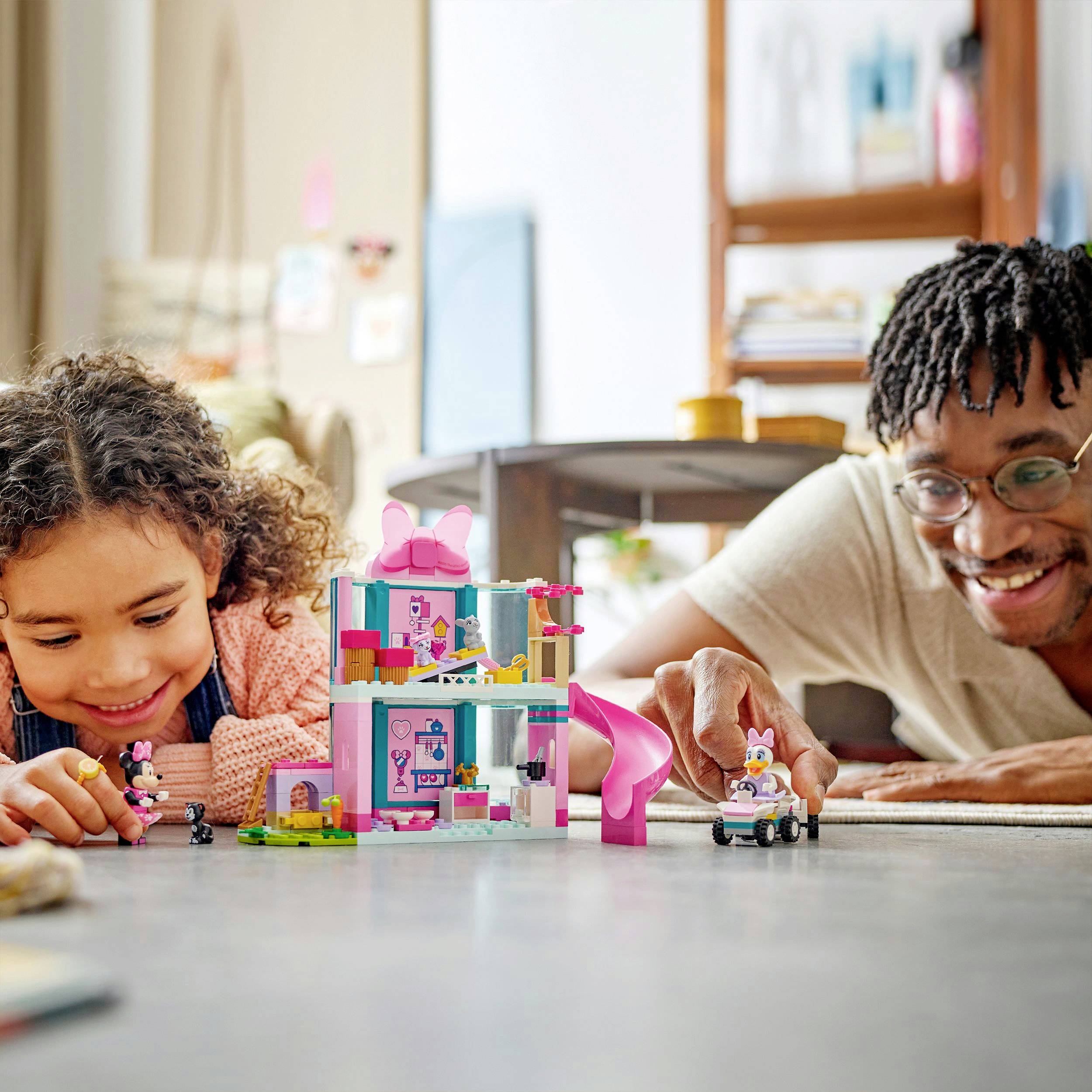 A child and an adult are playing on the floor with colorful building blocks and toy figures. Both are smiling and appear focused.