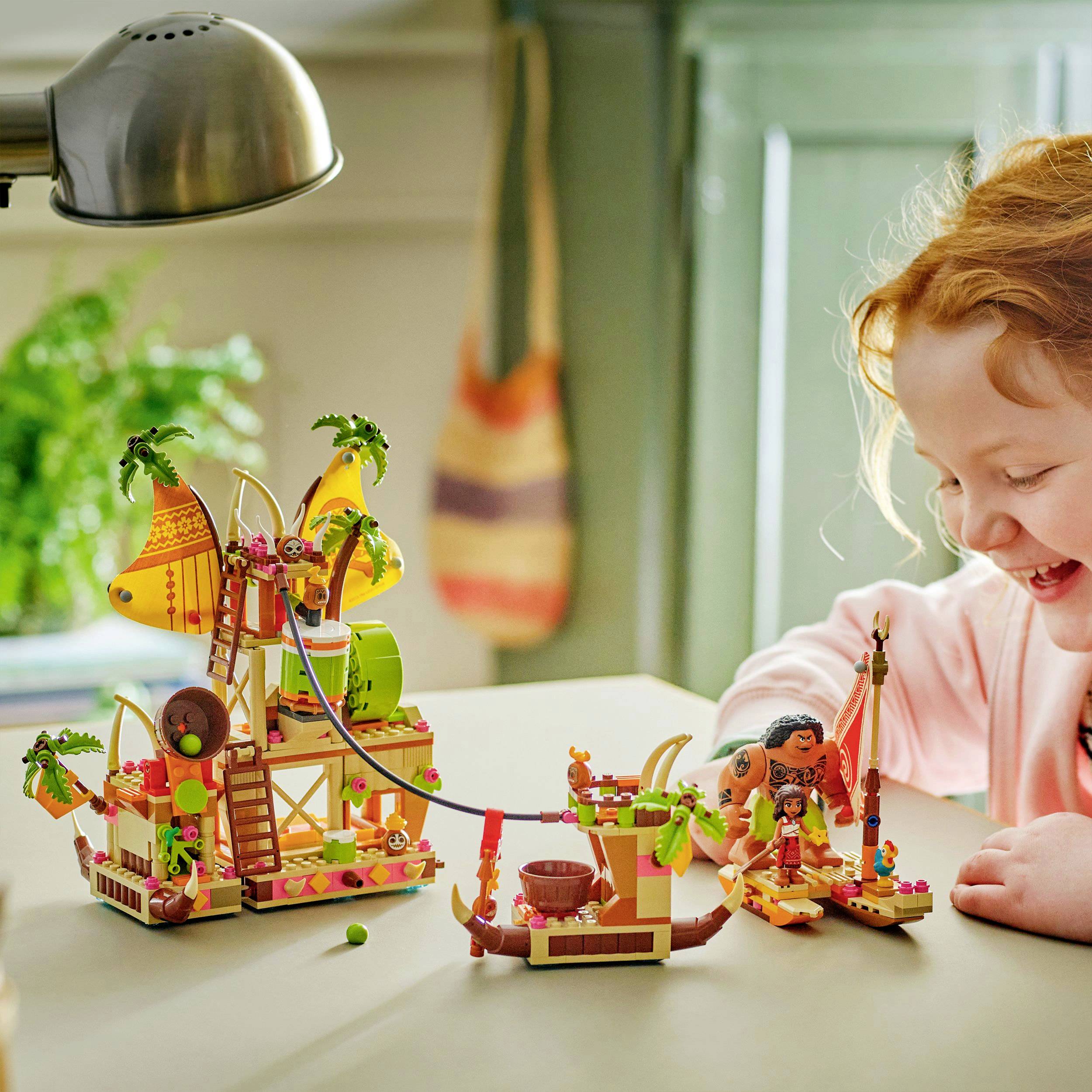 A child is joyfully playing with a colorful LEGO pirate ship set on a table. Blurred furniture can be seen in the background.