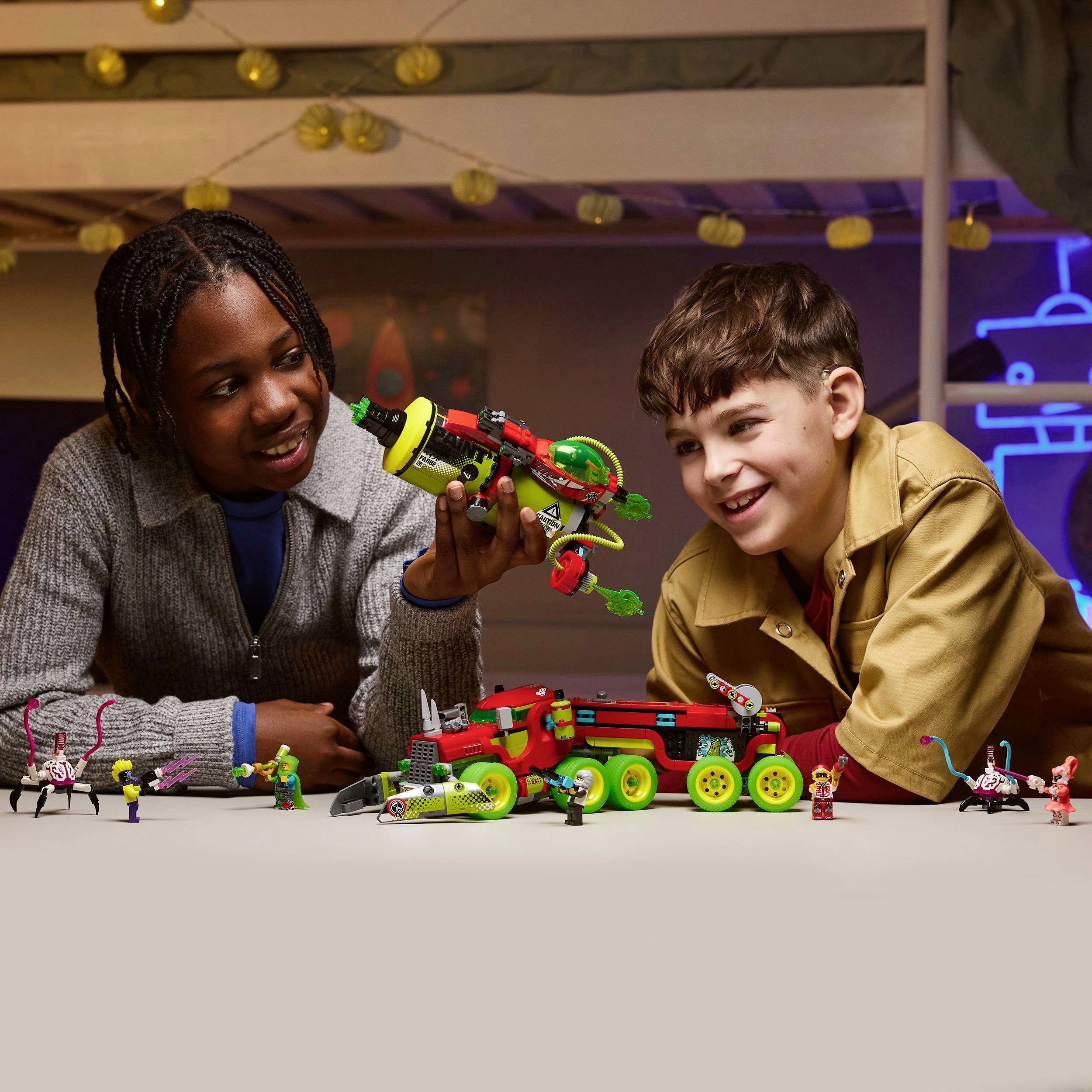 Two children are playing with colorful toys on a table. One is holding a model in their hand, while the other is laughing and watching.