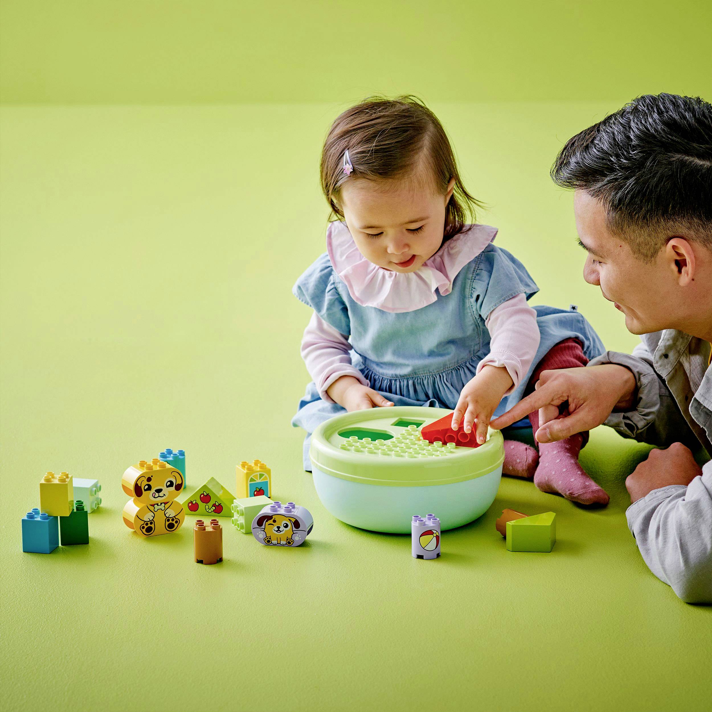 A small child is playing with building blocks while an adult sits nearby and smiles. Green walls can be seen in the background.