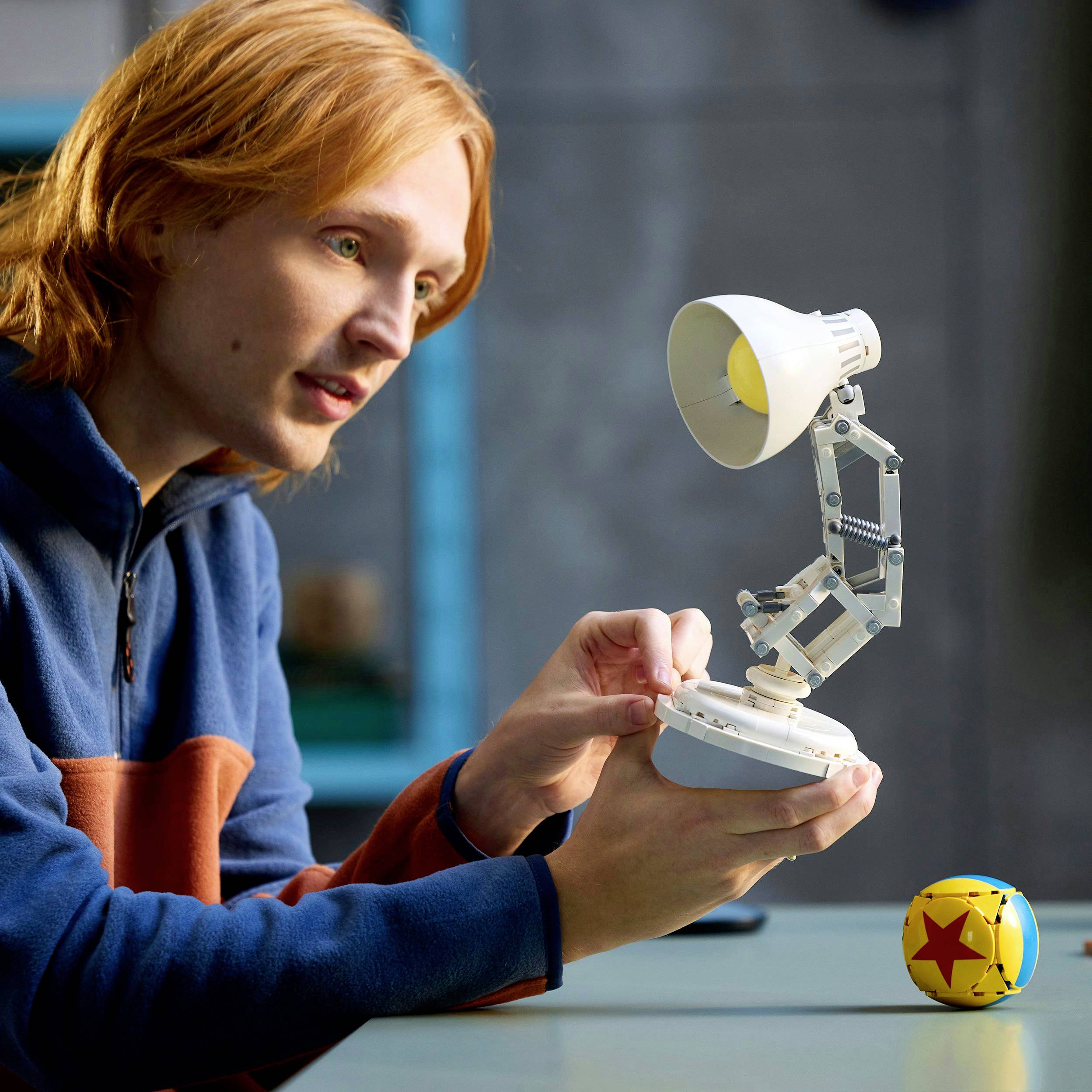A person holds a small lamp and operates a lever. Next to it lies a colorful ball. The scene suggests curiosity and discovery.