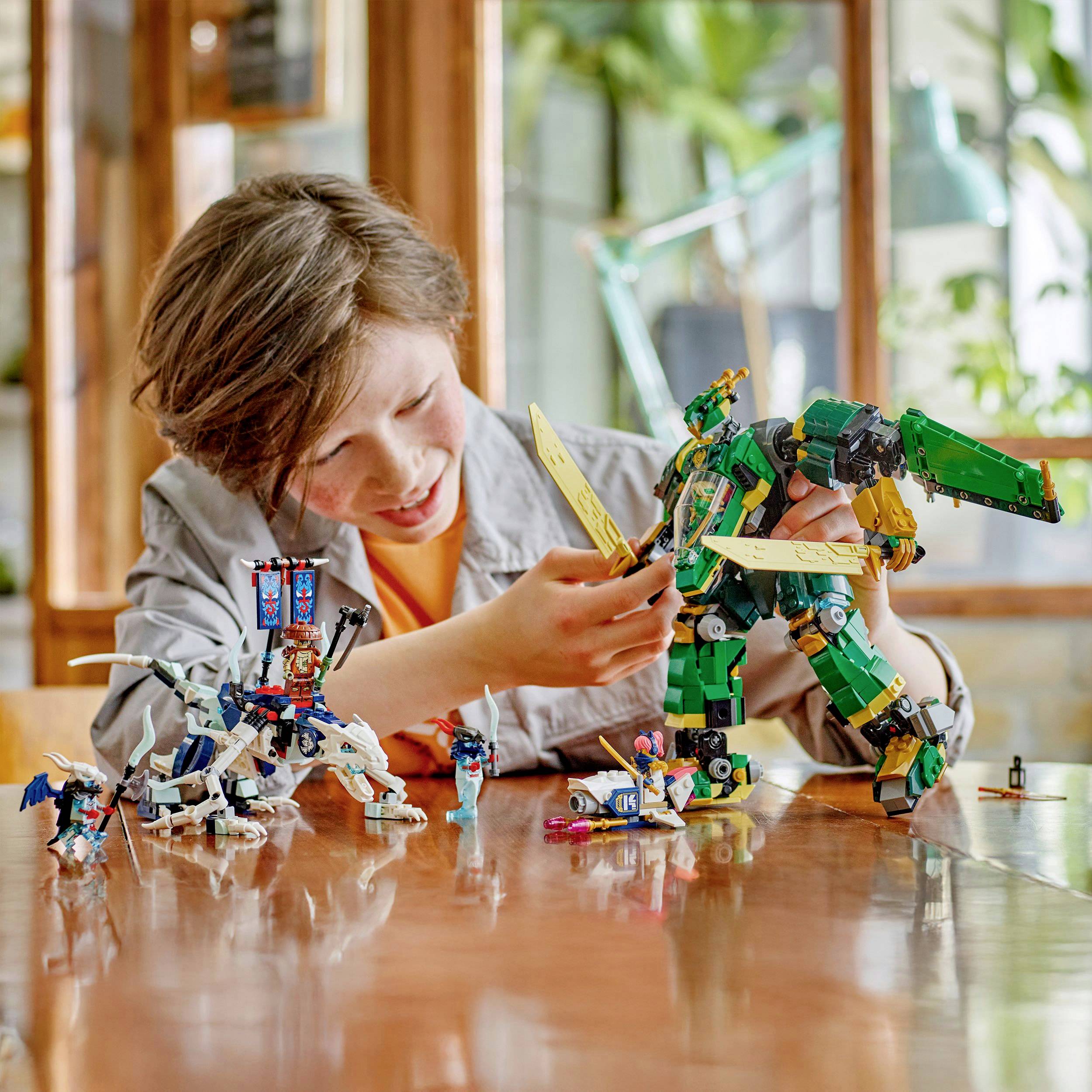 Boy plays enthusiastically with colorful robot models made from building blocks on a wooden table in a bright room with plants in the background.
