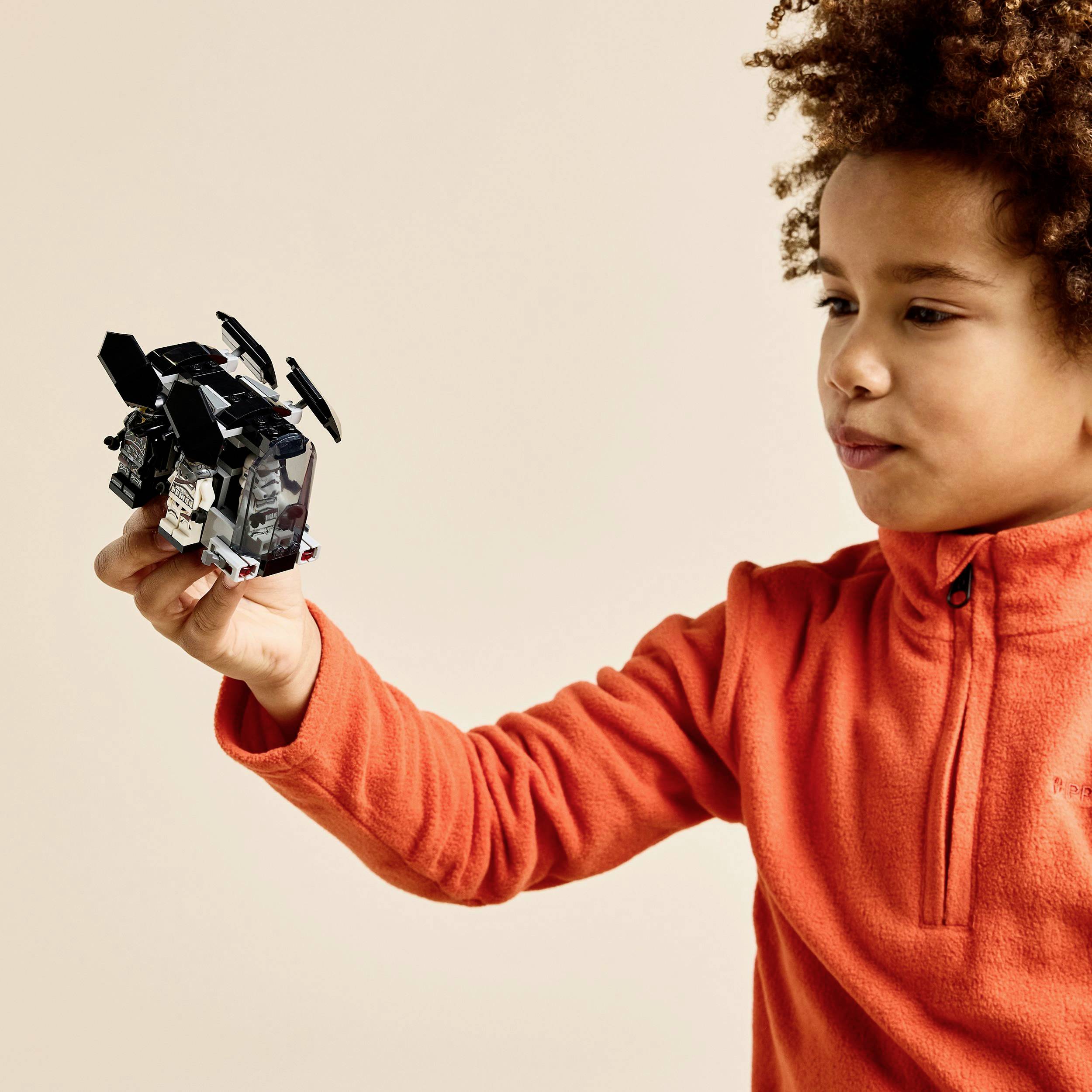 A child in an orange sweater is holding a toy robot in their hand and looking at it with interest.