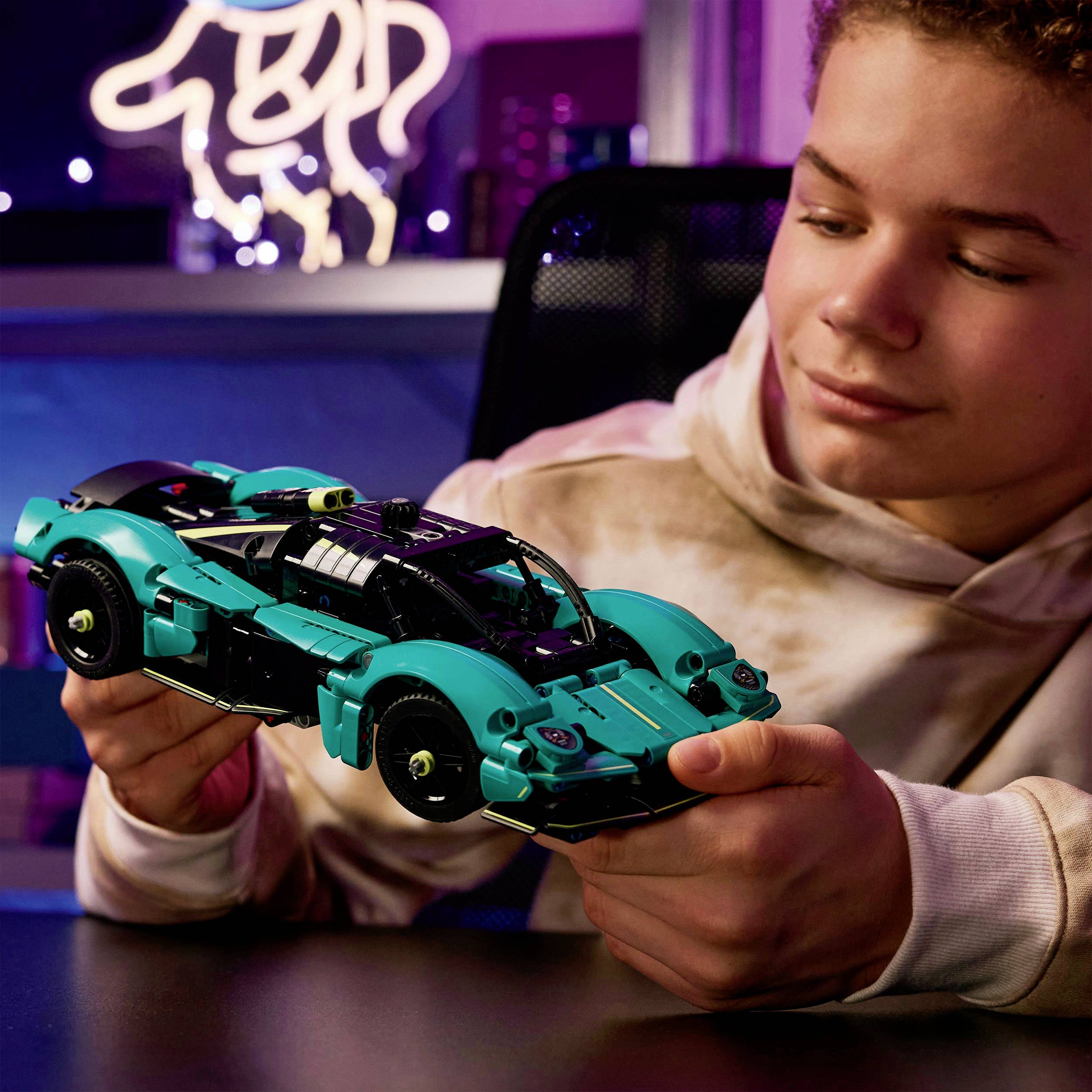 A child is looking at and holding a model car made of building blocks in a room with colorfully lit decoration in the background.