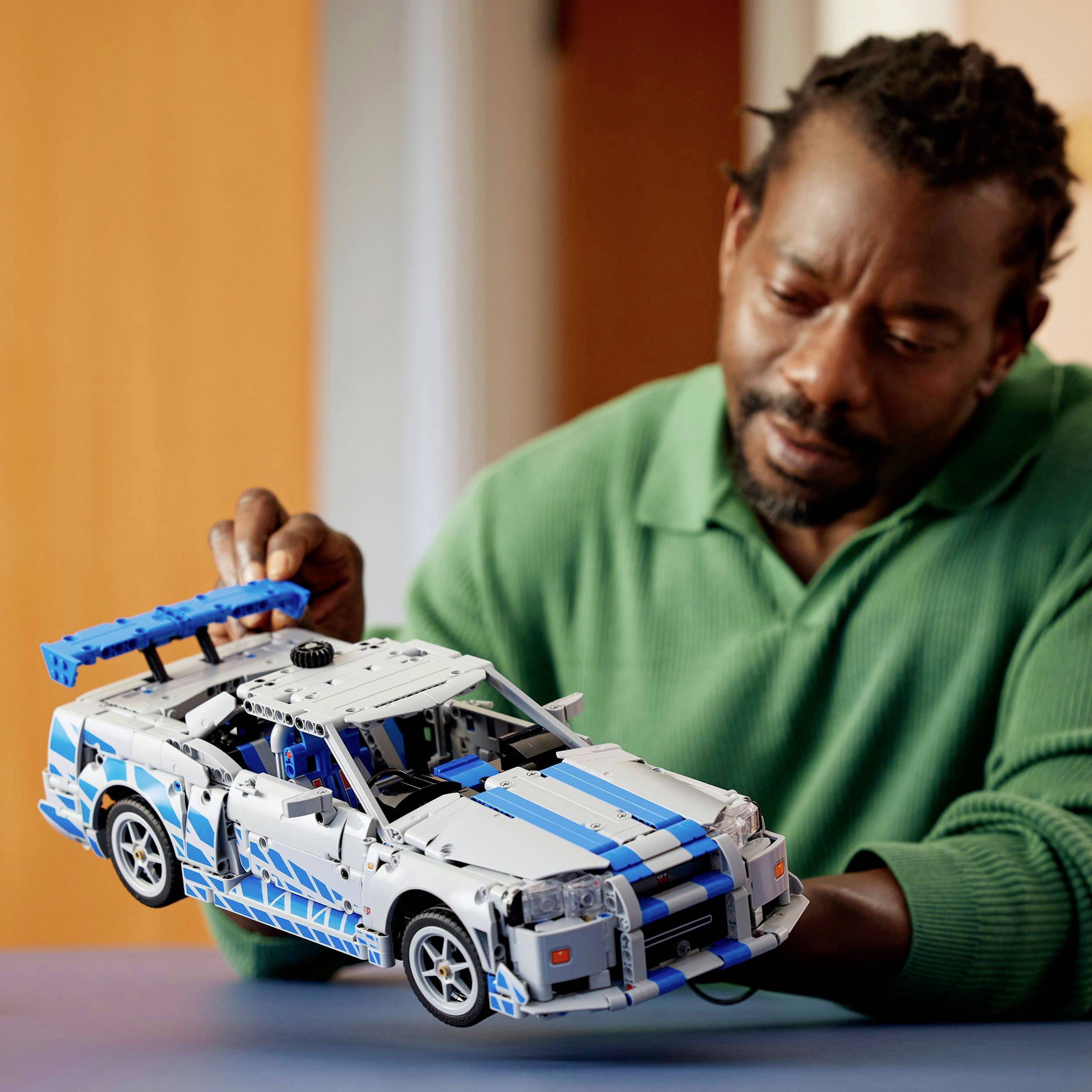 A man in a green sweater is looking at a model car made of building blocks that he is holding in his hand.