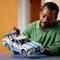A man in a green sweater is looking at a model car made of building blocks that he is holding in his hand.