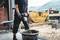 A worker is mixing concrete with a mixer on a construction site, with containers and lorries in the background.