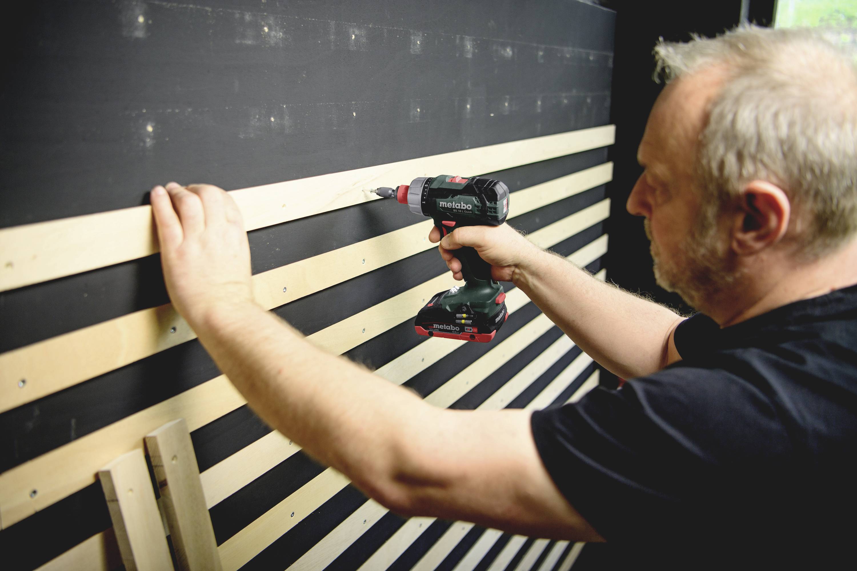 A man is drilling wooden battens into a wall. He is holding a cordless drill in his right hand and aligning the battens with his left hand.
