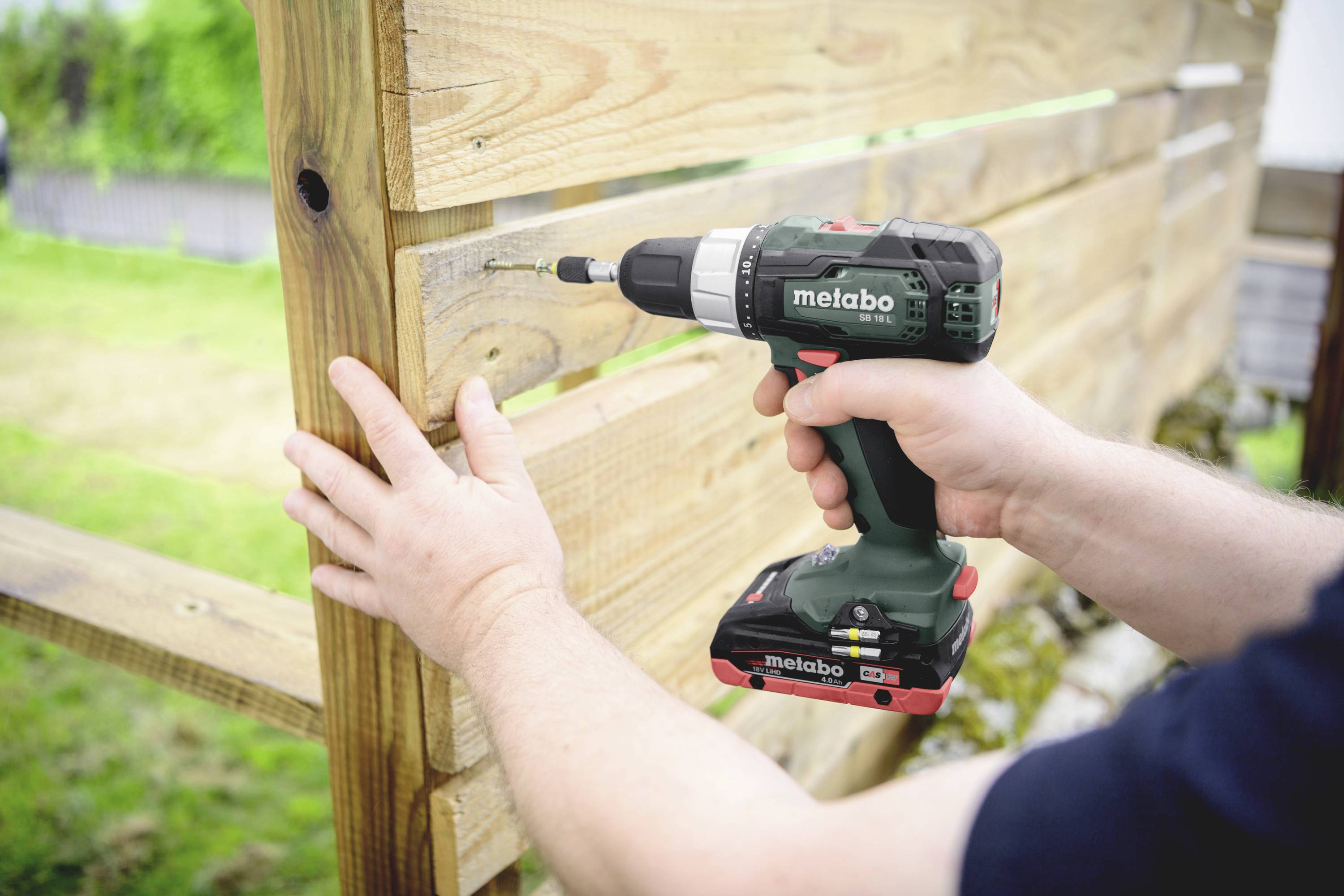 A person is installing a wooden wall outdoors using a cordless drill. Hands are holding the wood for stability.