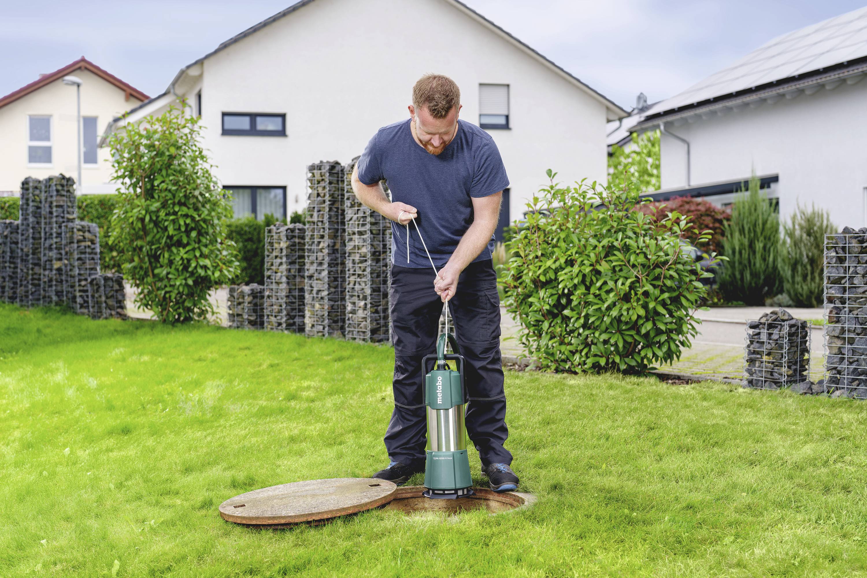 A man lifts a green pump from a circular well in a verdant lawn in front of a white house.