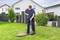 A man lifts a green pump from a circular well in a verdant lawn in front of a white house.