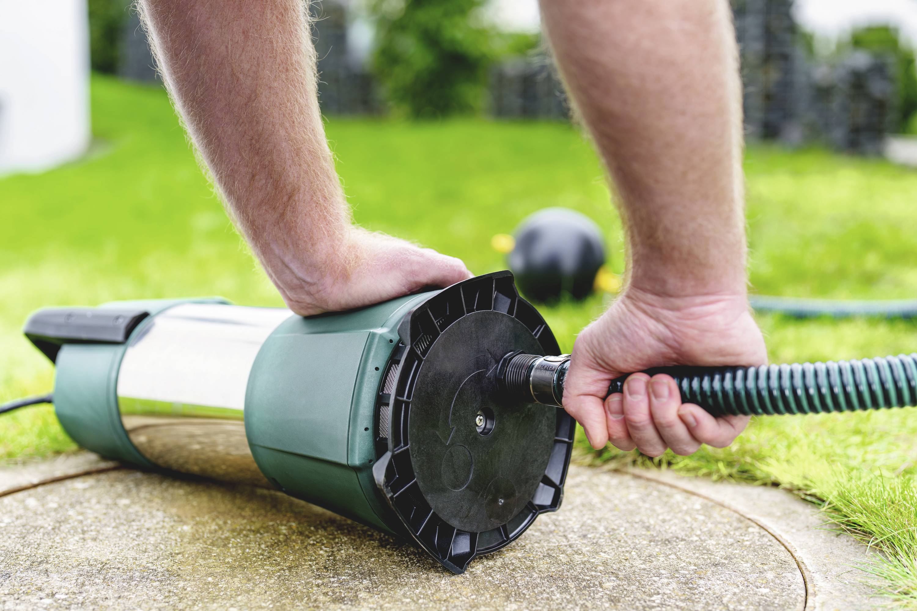 A person is attaching a green garden hose to a grey water pump in the garden to pump water.