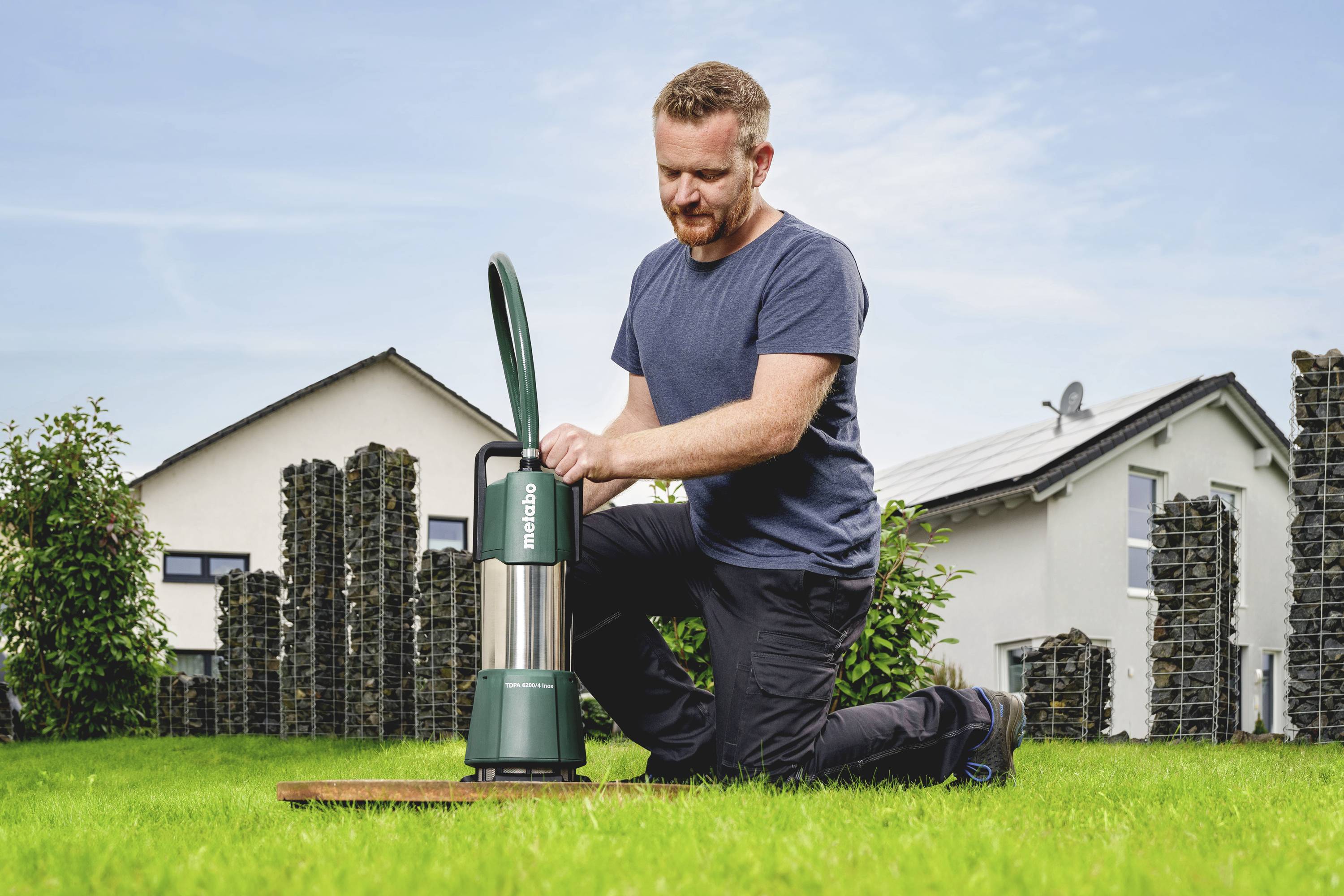A person is kneeling on a lawn and holding a green submersible pump. A house and some shrubs can be seen in the background.