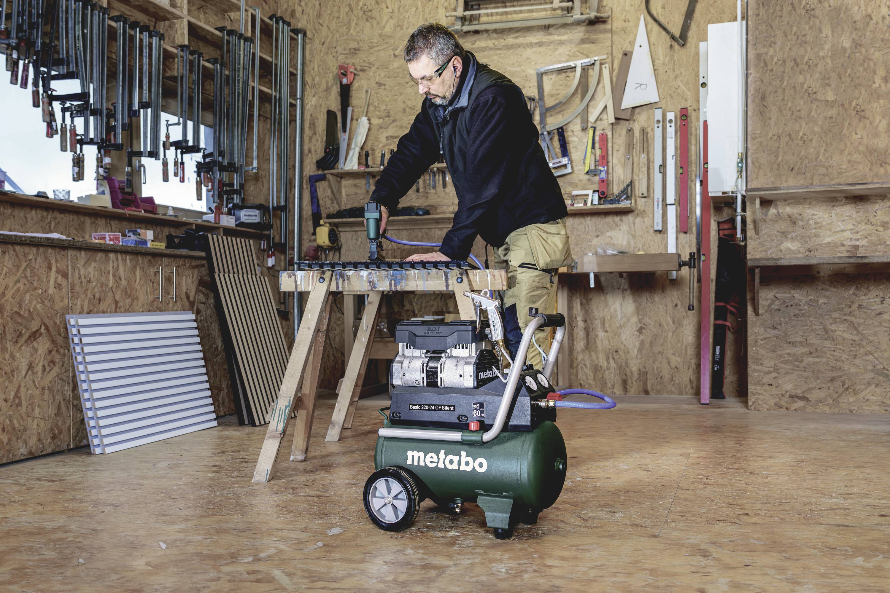 A man is working in a workshop with wooden walls. In front of him stands a portable air compressor on the floor.