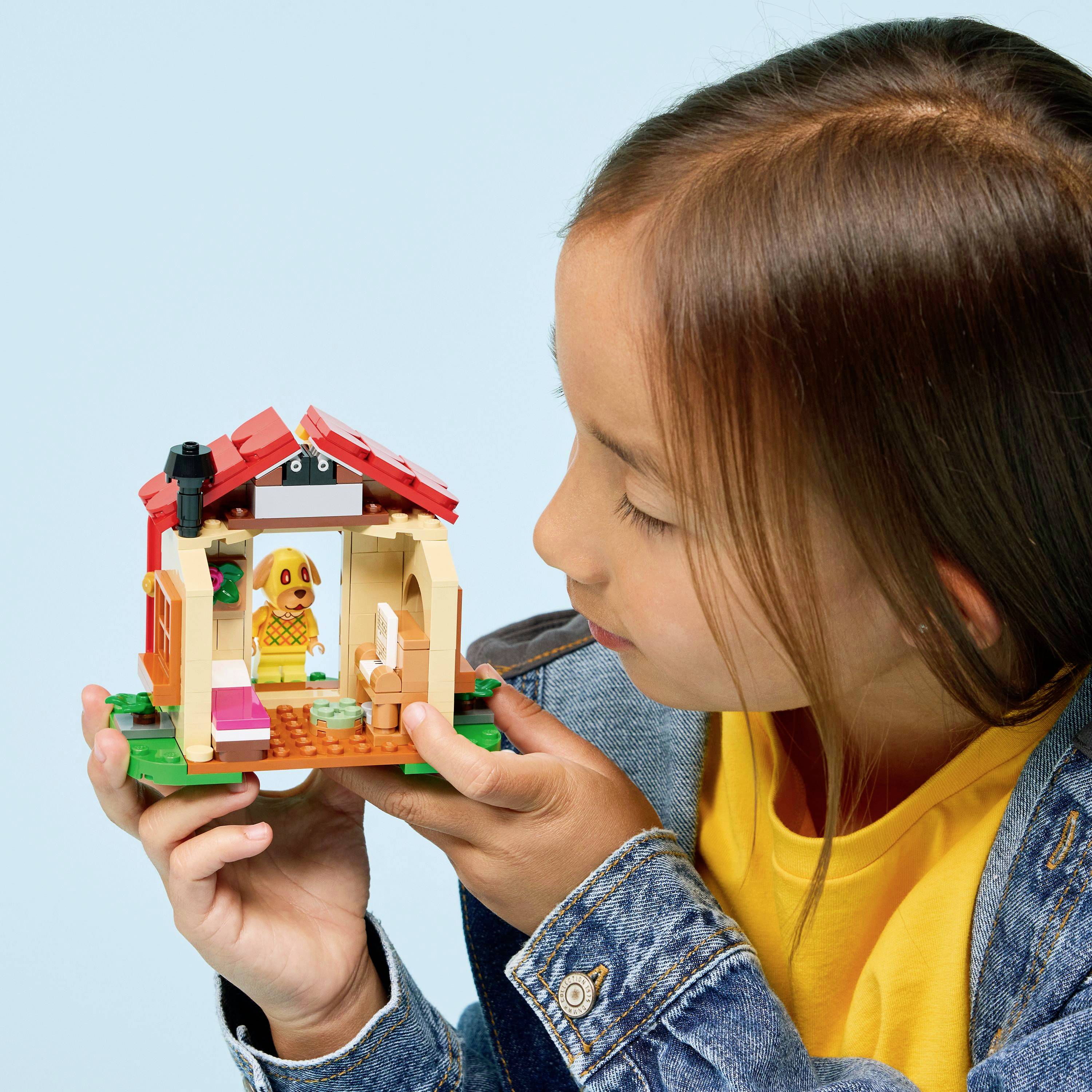 A child in a denim jacket smiles while holding a small, colorful toy house with an open roof, revealing miniature furniture inside.