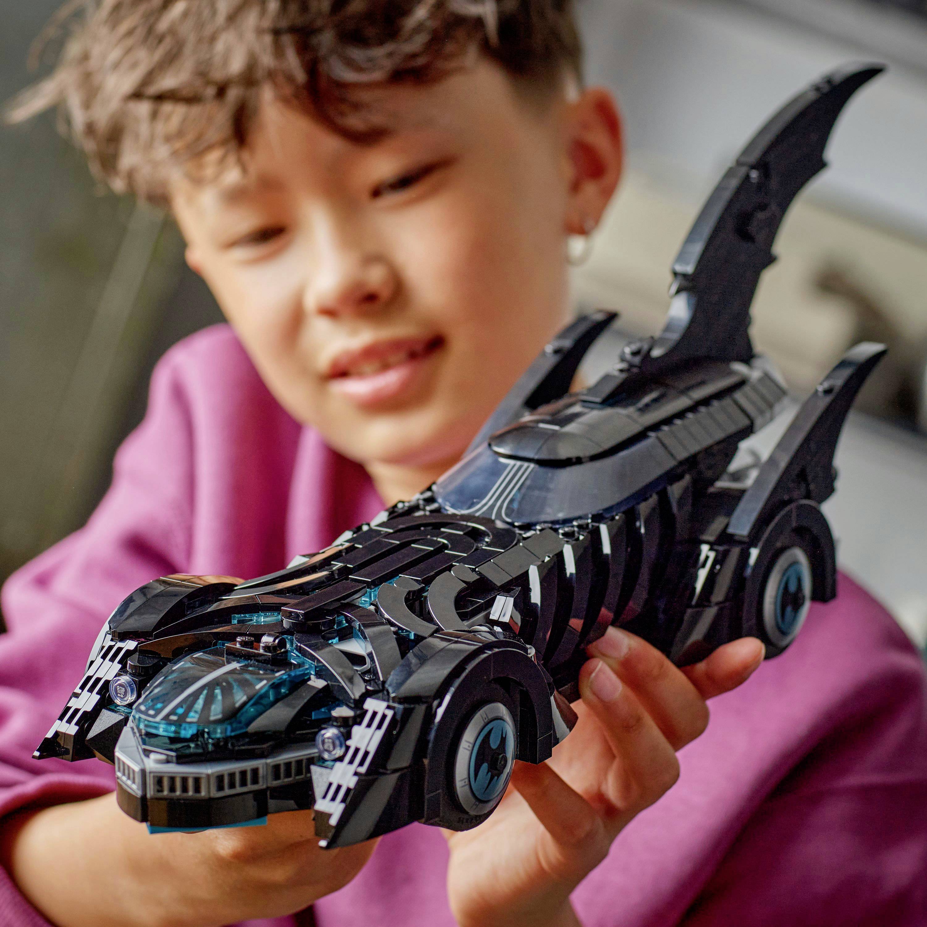 A person holding a detailed model of a black, futuristic car with sharp fins, resembling a popular superhero vehicle, indoors.
