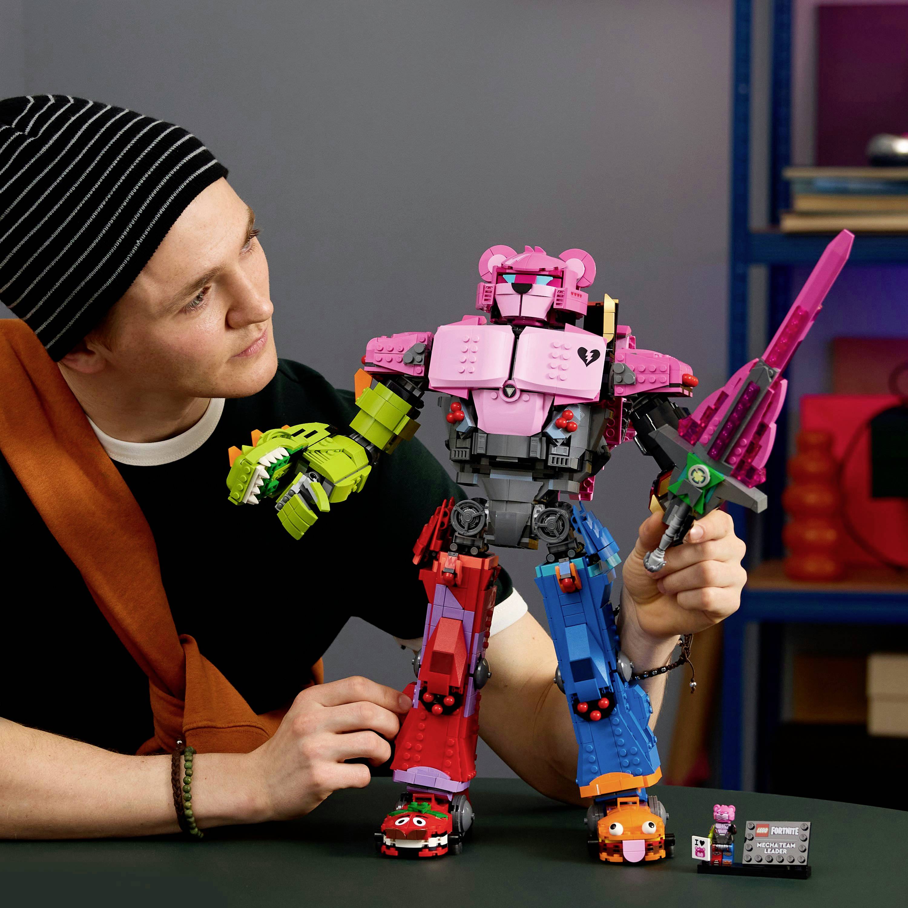 A person in a striped beanie admires a colorful, block-style robot with animal features, holding a pink sword and a toy truck.