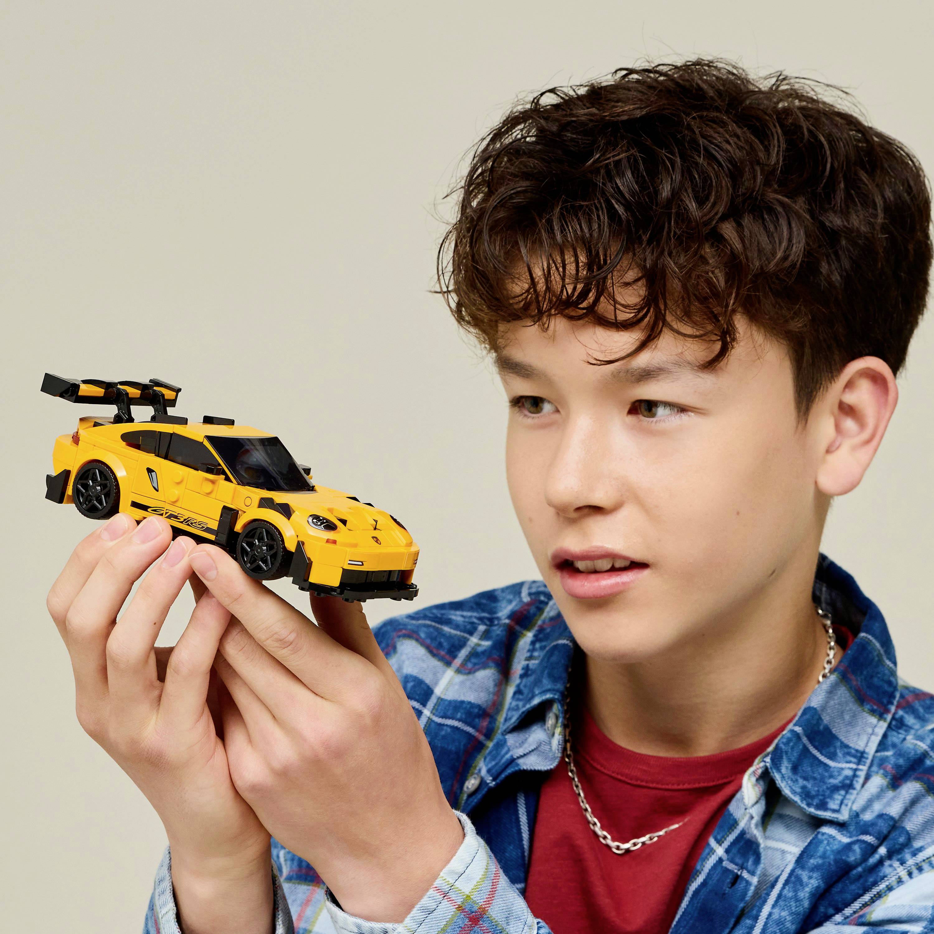 A young person holds a yellow toy car model, looking at it with interest. They wear a blue checked shirt and red t-shirt.
