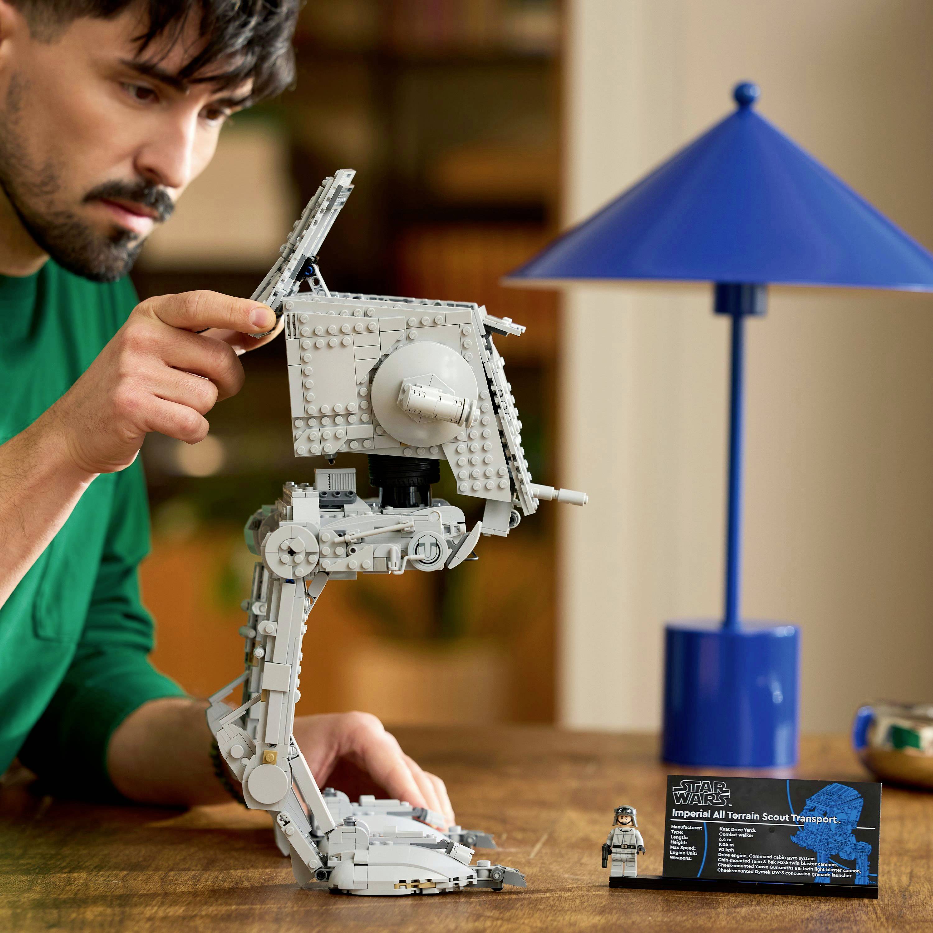 A man assembles a detailed model of an Imperial All Terrain Scout Transport on a table, with a blue lamp in the background.