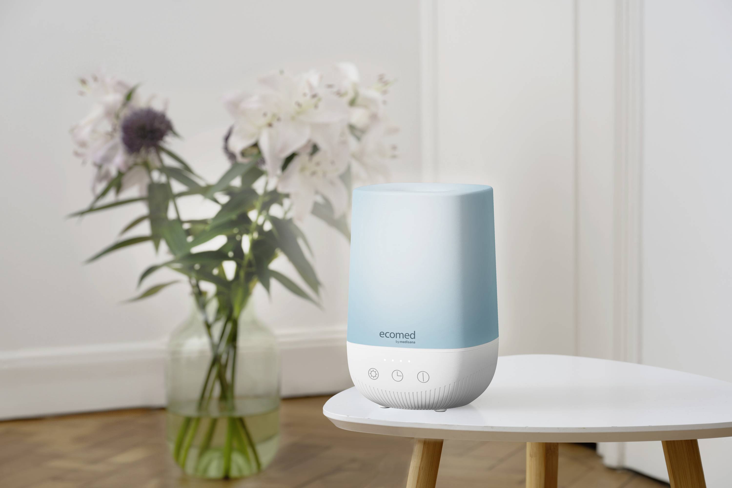 A blue Ecomed air humidifier sits on a white table, with a vase of flowers in the background.