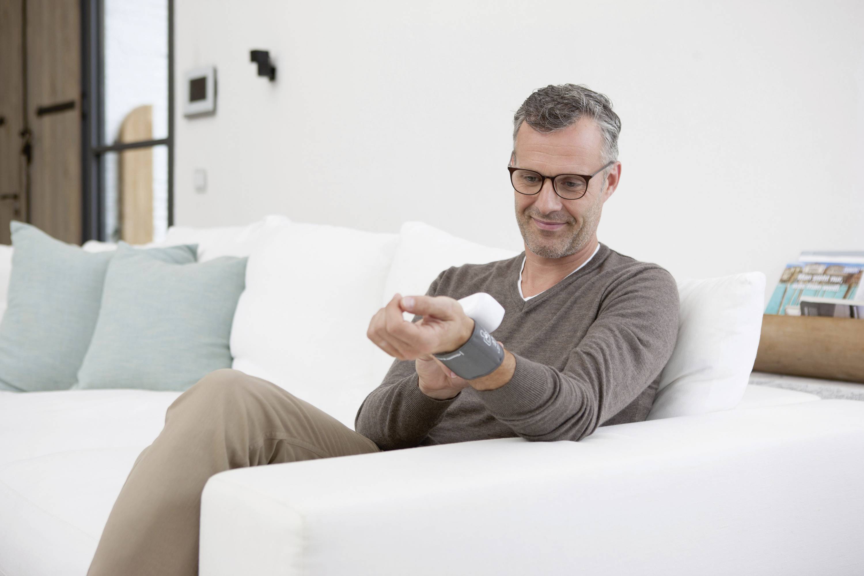 A man checks his blood pressure using a wrist blood pressure monitor while sitting on a sofa.