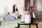 An air purifier stands on a table next to a vase with lilac orchids. In the background, a blurry person sits on a sofa.