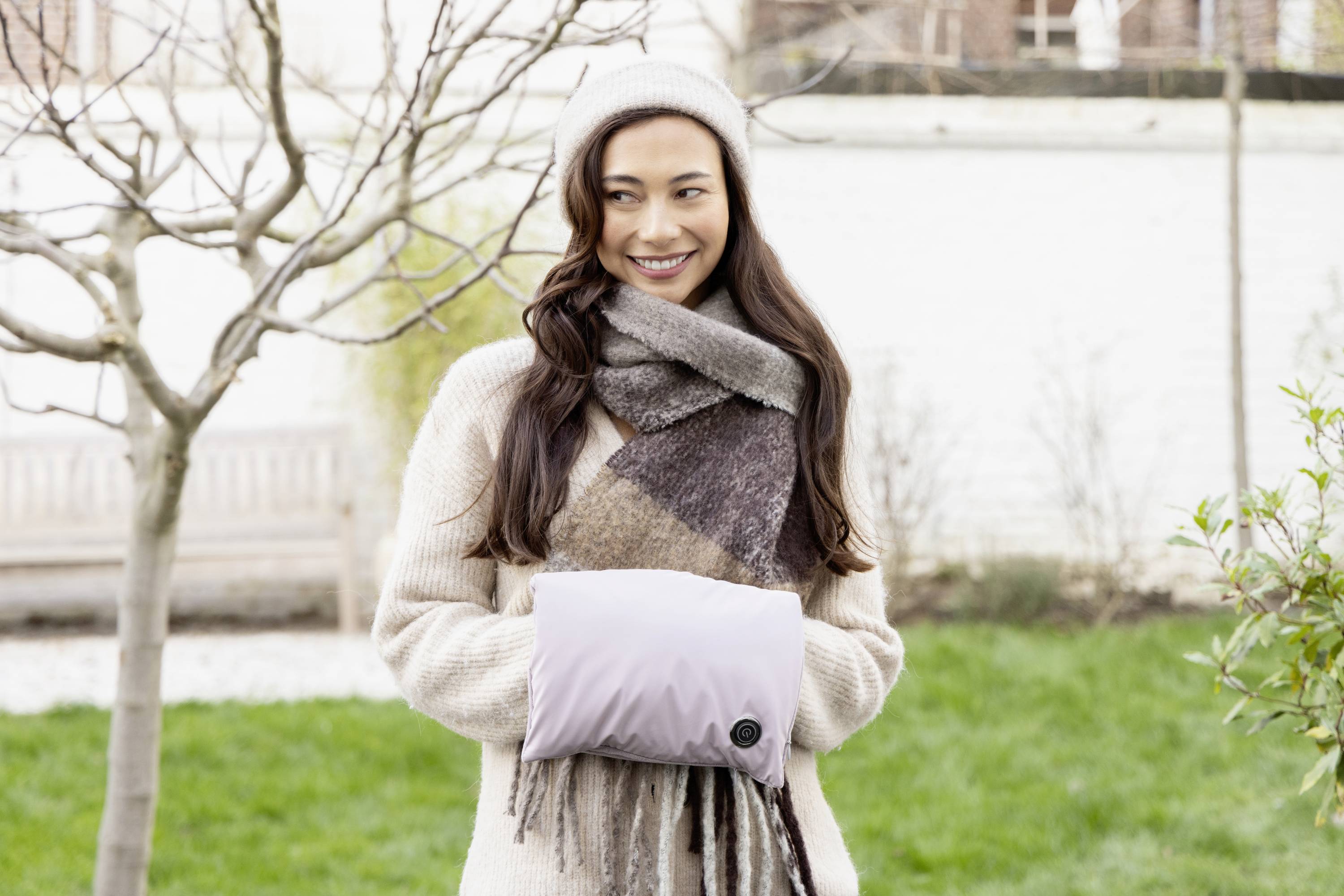 A smiling woman stands outdoors, wearing warm clothing, including a scarf and hat, and is holding a cushion in front of her. Trees and a lawn are visible in the background.