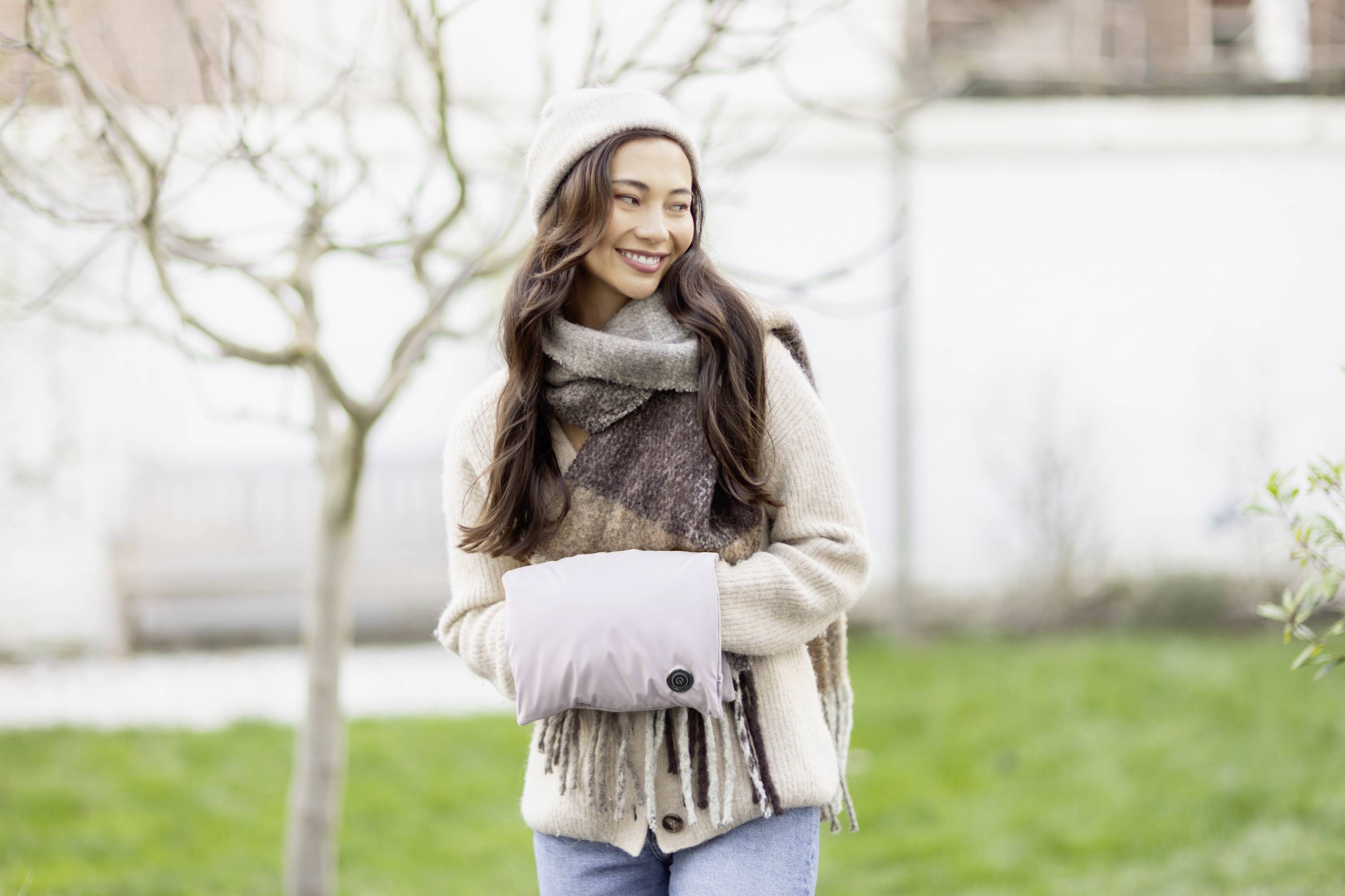 Woman wearing a scarf and hat smiles outside in the garden. She is wearing a beige jacket and holding a muff-like hand warmer. The sky is slightly overcast.