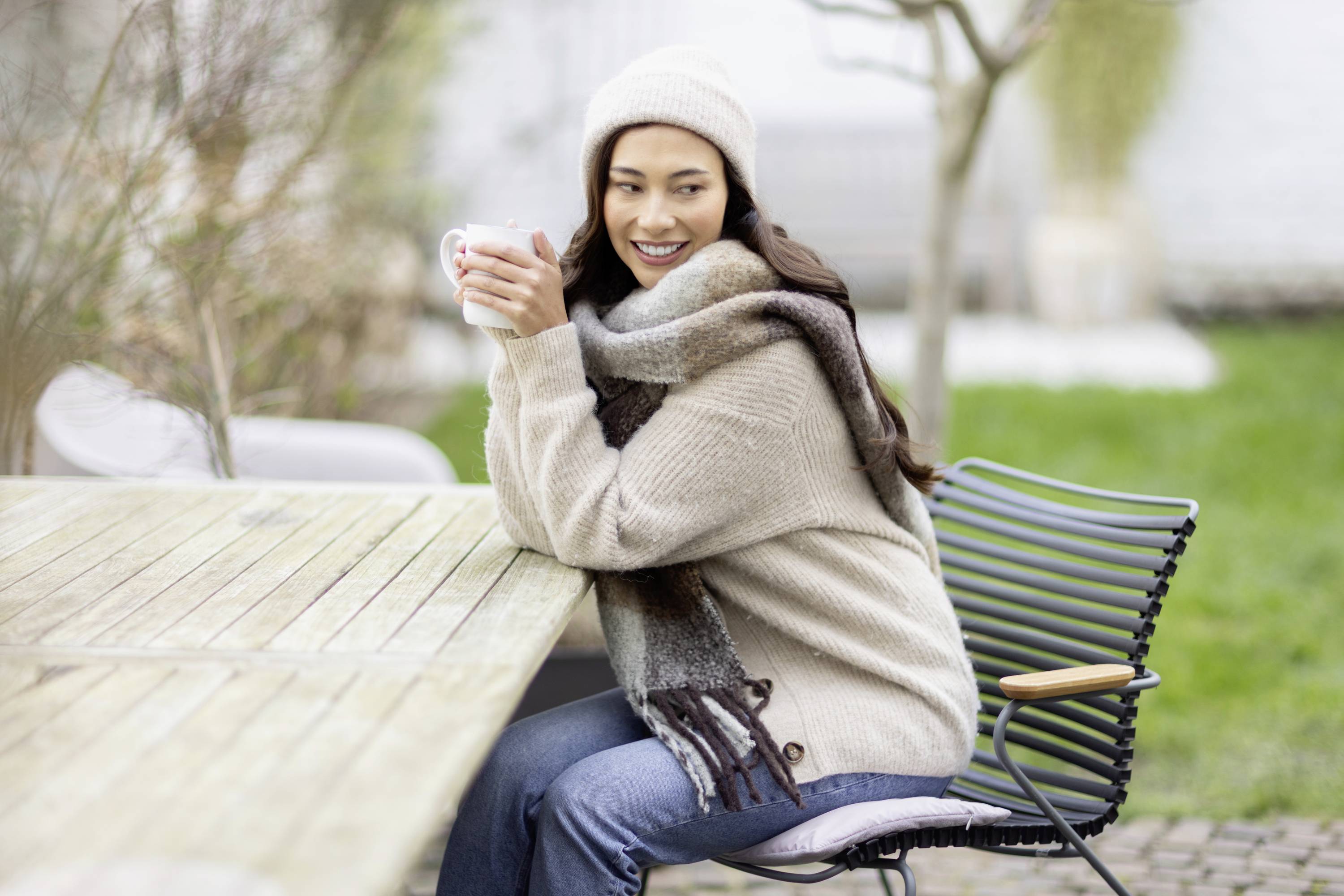 A smiling person is sitting outdoors at a wooden table, holding a mug. They are wearing a warm jumper, scarf and woolly hat.