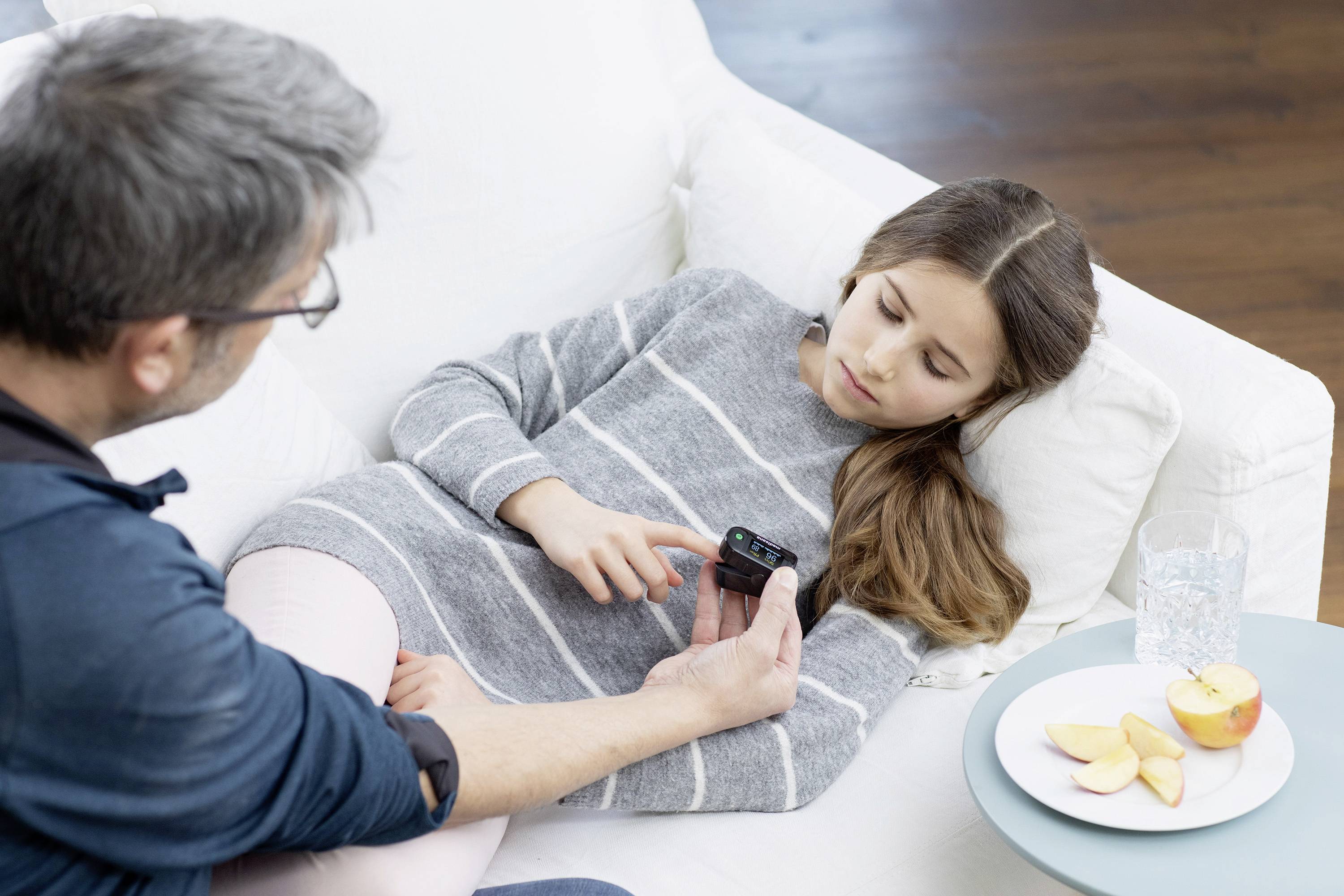 A man is showing a girl on a sofa a medical device. Next to her is a plate with apple slices and a glass of water.