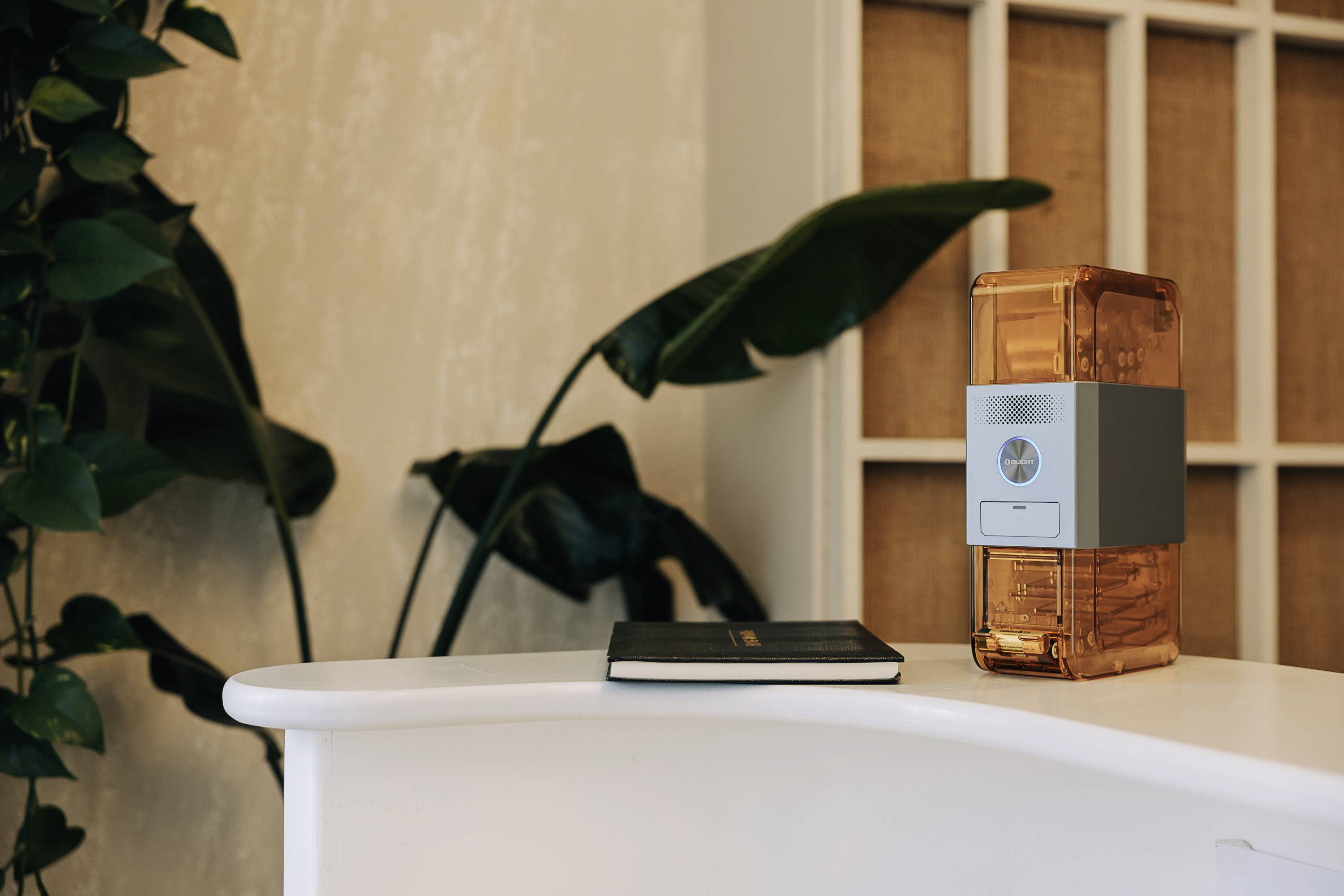A small golden device sits on a white table next to a black notebook. Plants are visible in the background.