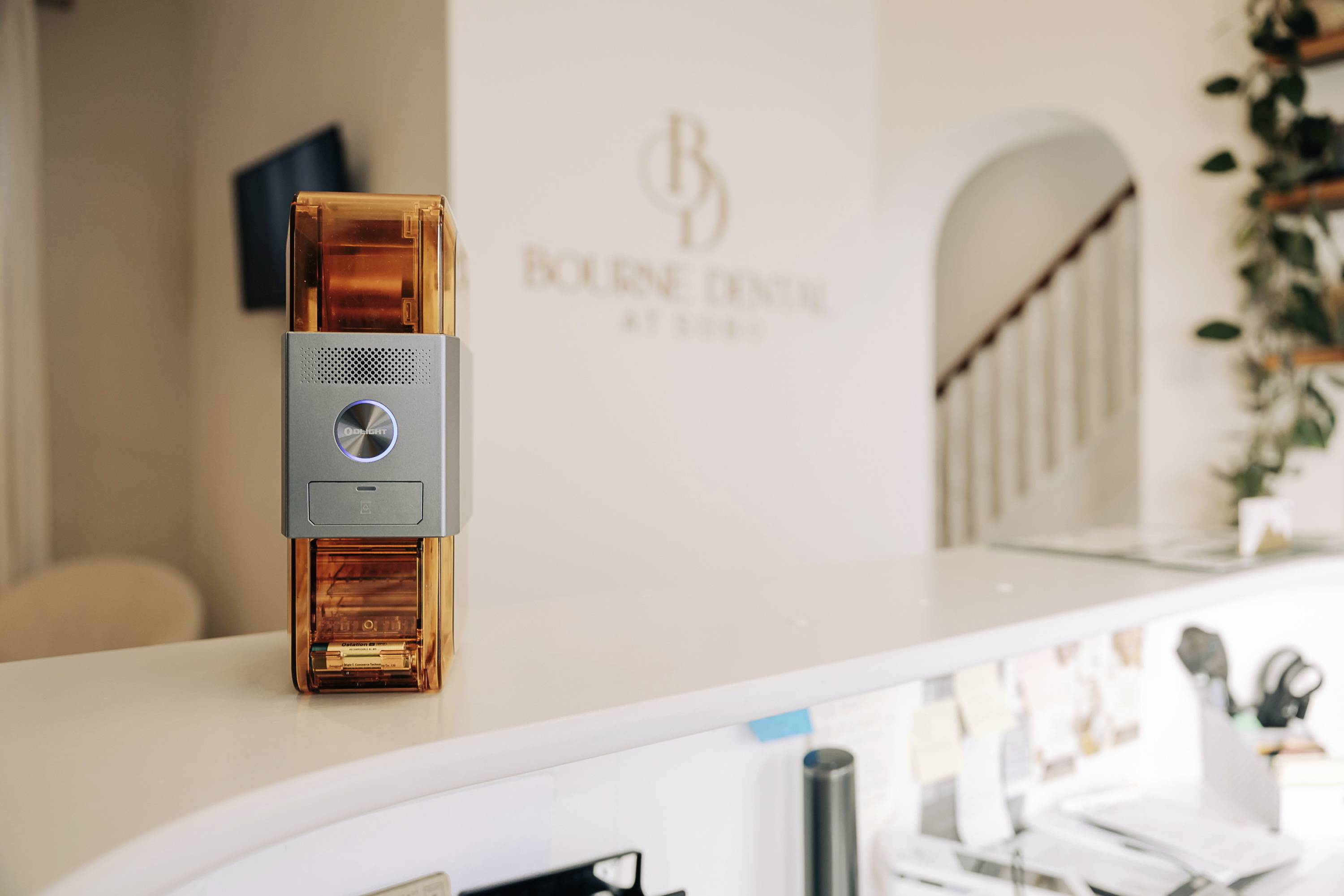 A transparent orange dental scanner sits on a reception desk in a dental practice, with a logo visible in the background.