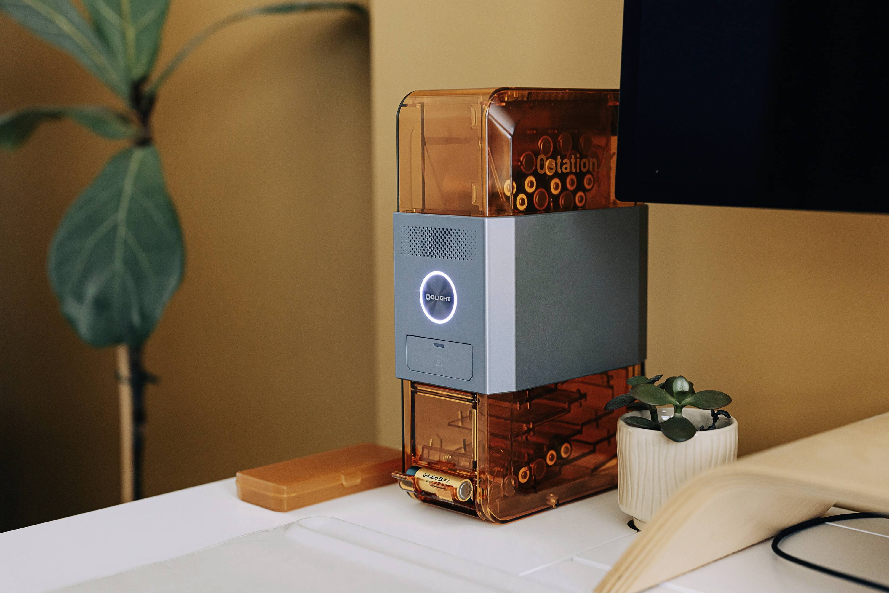 3D printer on a table next to a monitor and a plant. The printer is orange-transparent and displays 'Cetation' on the front.