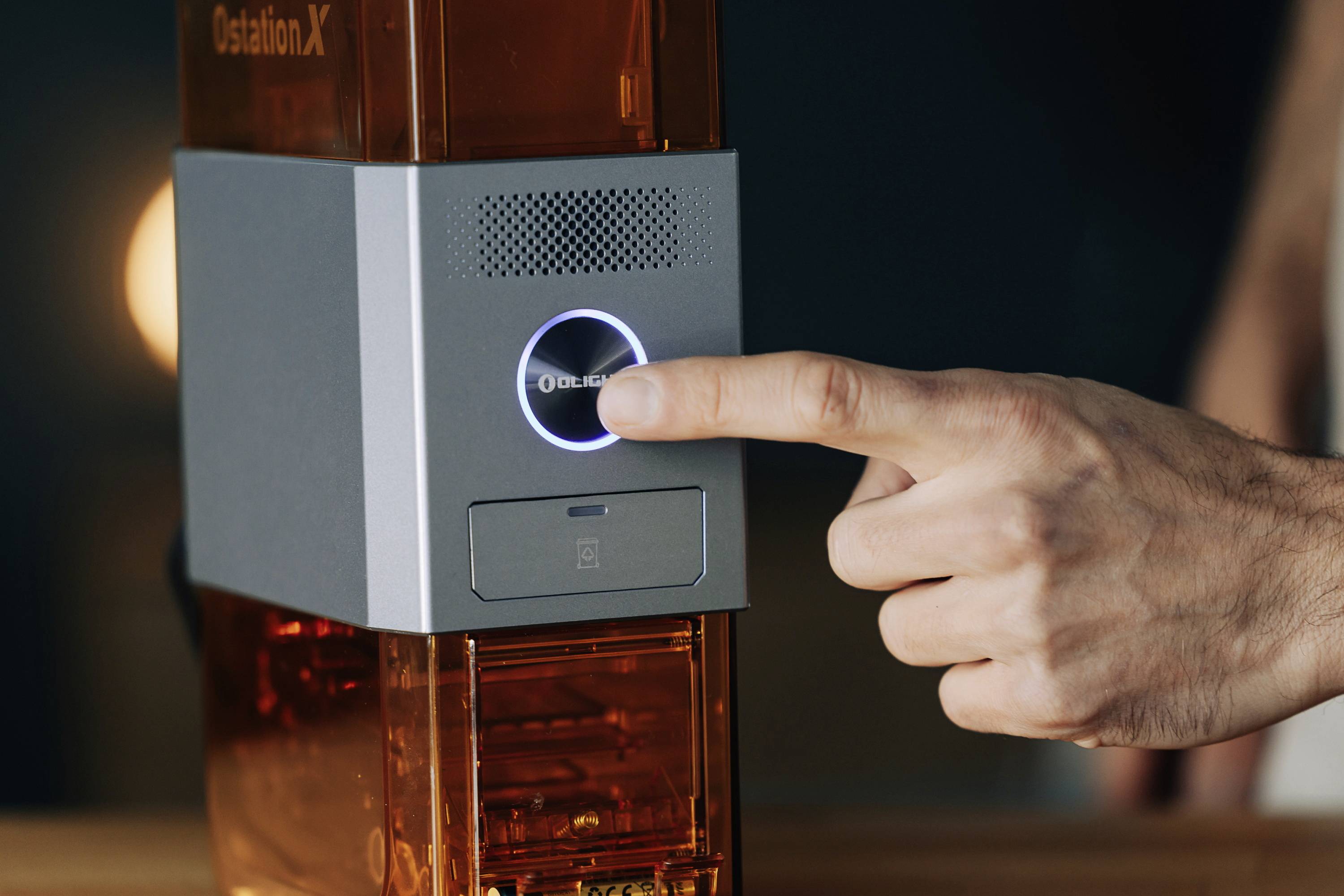 A hand presses the illuminated 'Start' button of a 3D printer. Blurred lights are visible in the background.