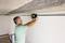 A man in a green shirt is using a measuring device on a white wall, apparently conducting precision measurements for a construction project.