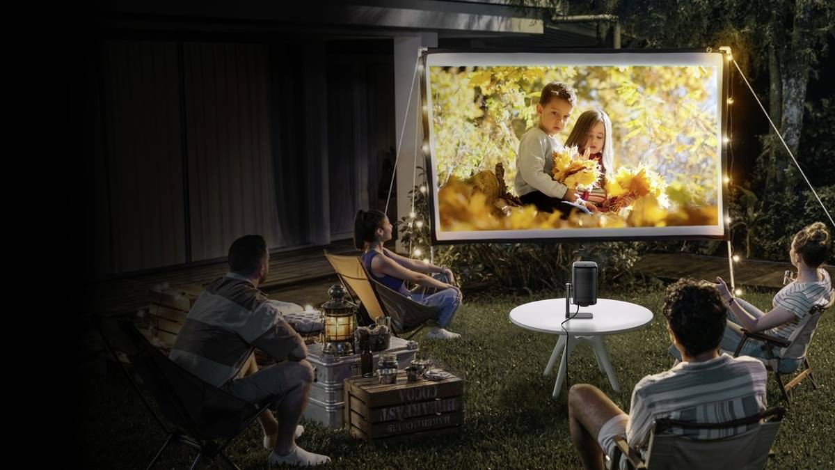 A group of three adults and two children are watching a film on a screen in the garden in the evening.