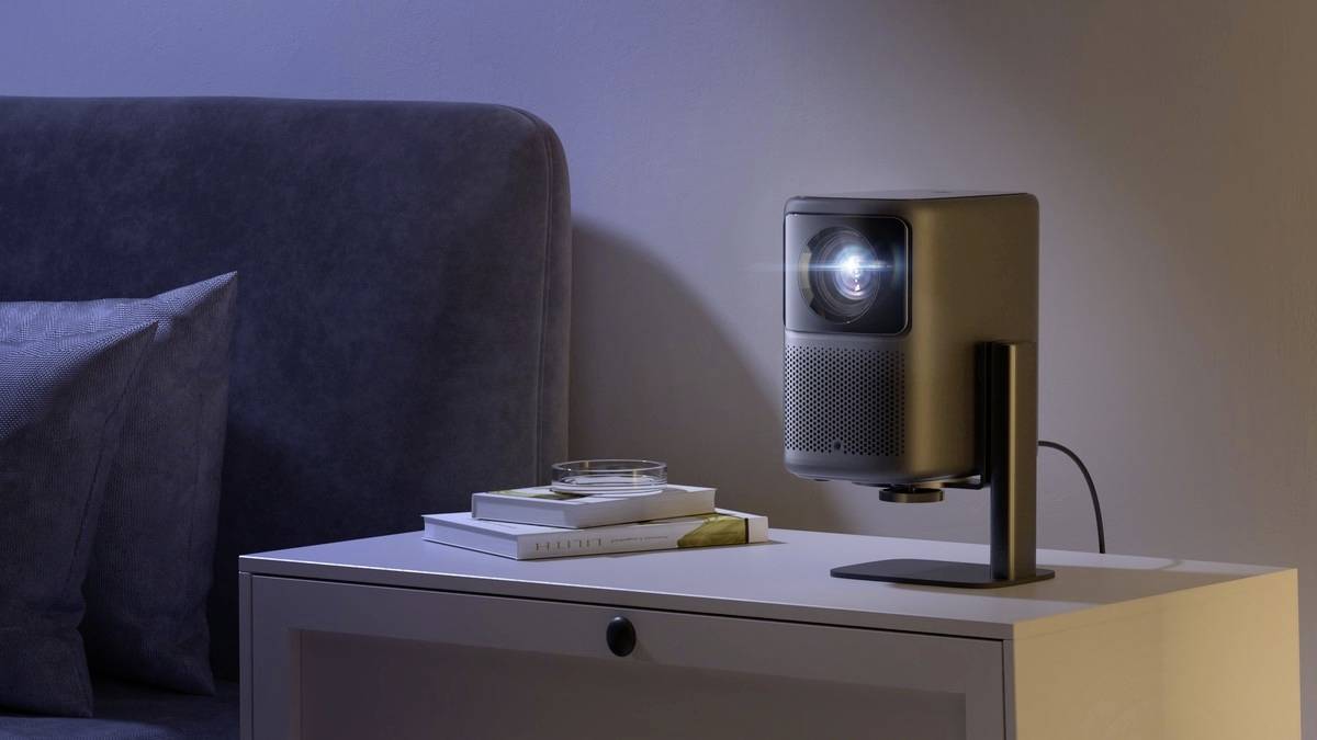 A compact projector sits on a bedside table next to a bed. The projector is displaying an image on the wall. Books are lying nearby.