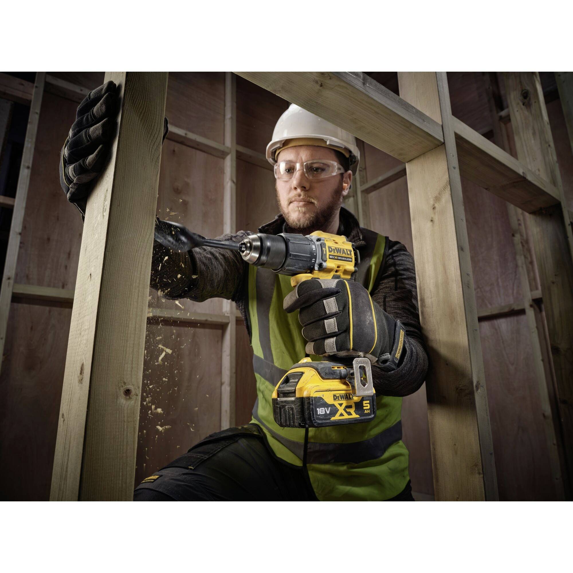 A person wearing safety glasses and a hard hat is drilling into a wooden frame with a cordless drill. Wood shavings are flying, and construction worker attire is visible.