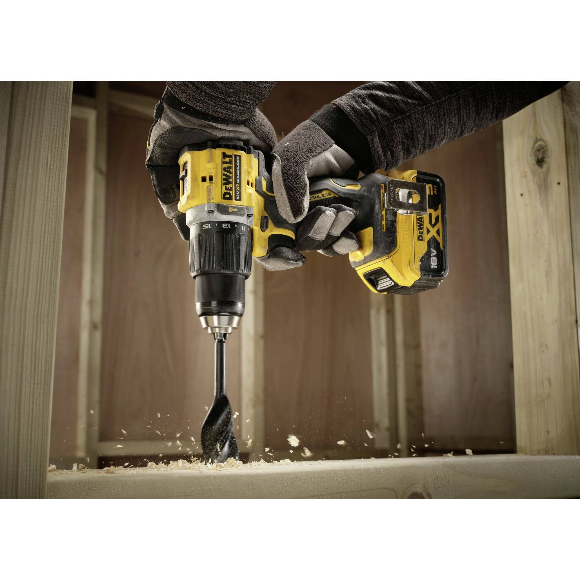 A person is drilling a hole in a piece of wood using a yellow, cordless drill, with wood shavings flying around.