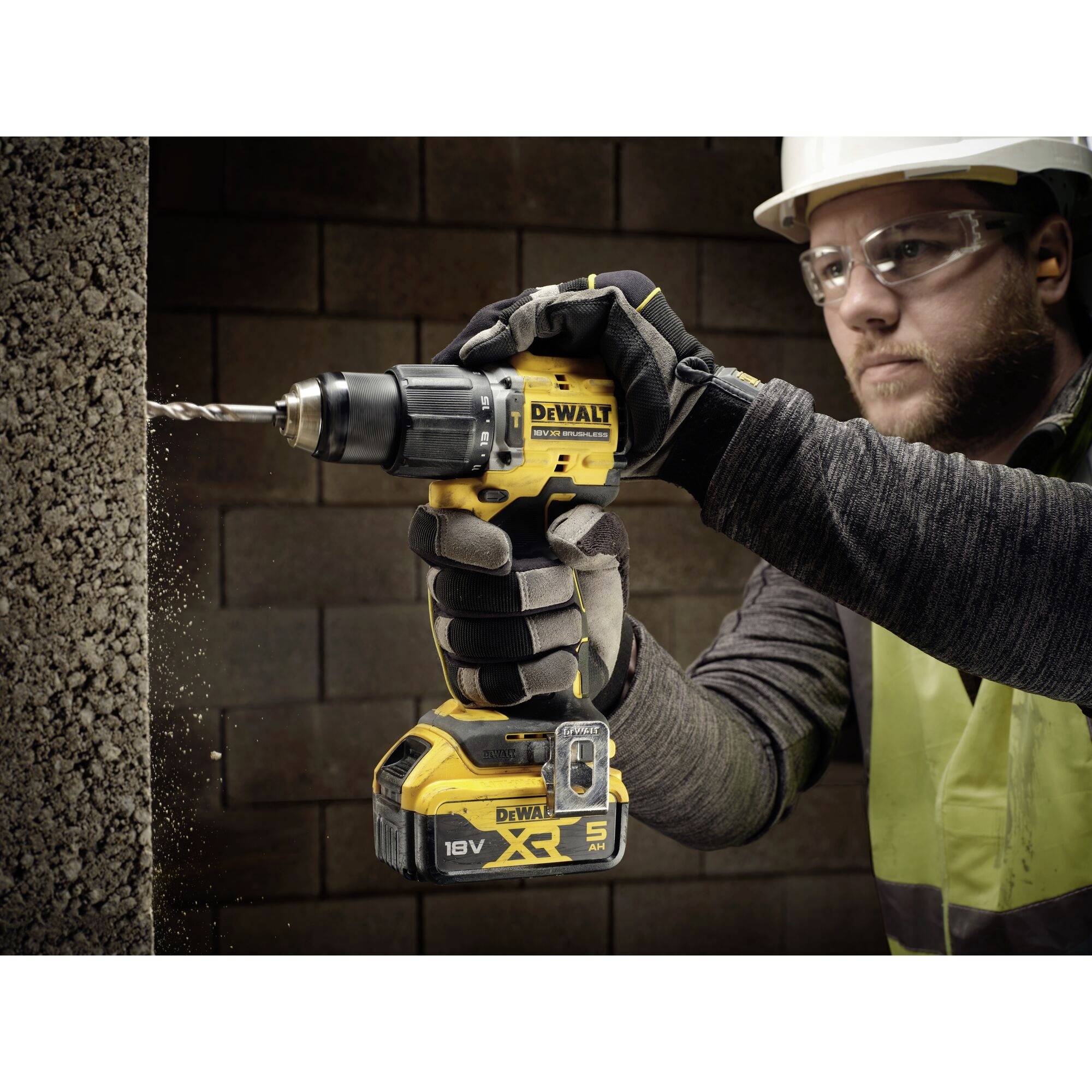 A person wearing safety glasses and gloves is using a yellow cordless drill to bore a hole in a concrete wall.