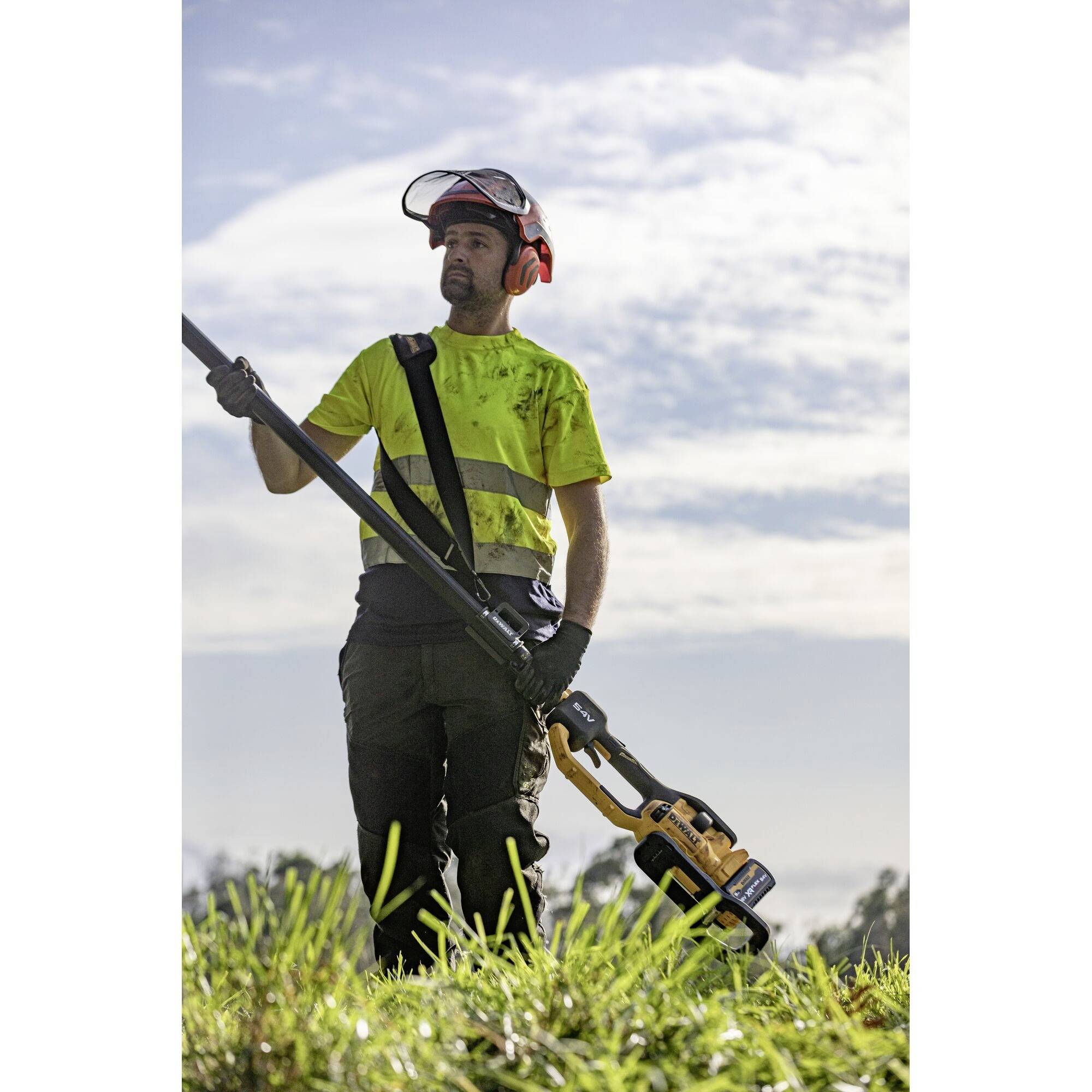 A person wearing a safety helmet and high-visibility vest is cutting grass with a strimmer on a field, with bright clouds in the sky in the background.