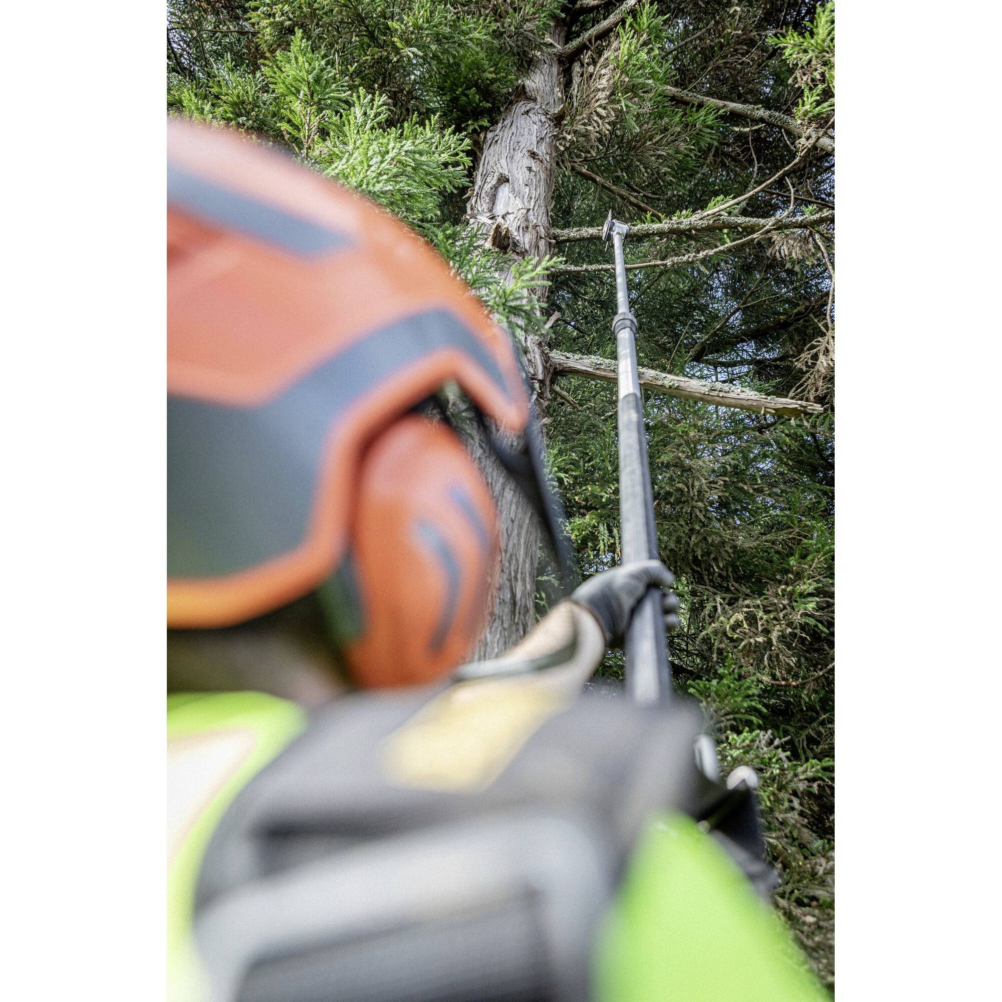 A person wearing a helmet and protective clothing is cutting branches from a tree using a pruning saw.