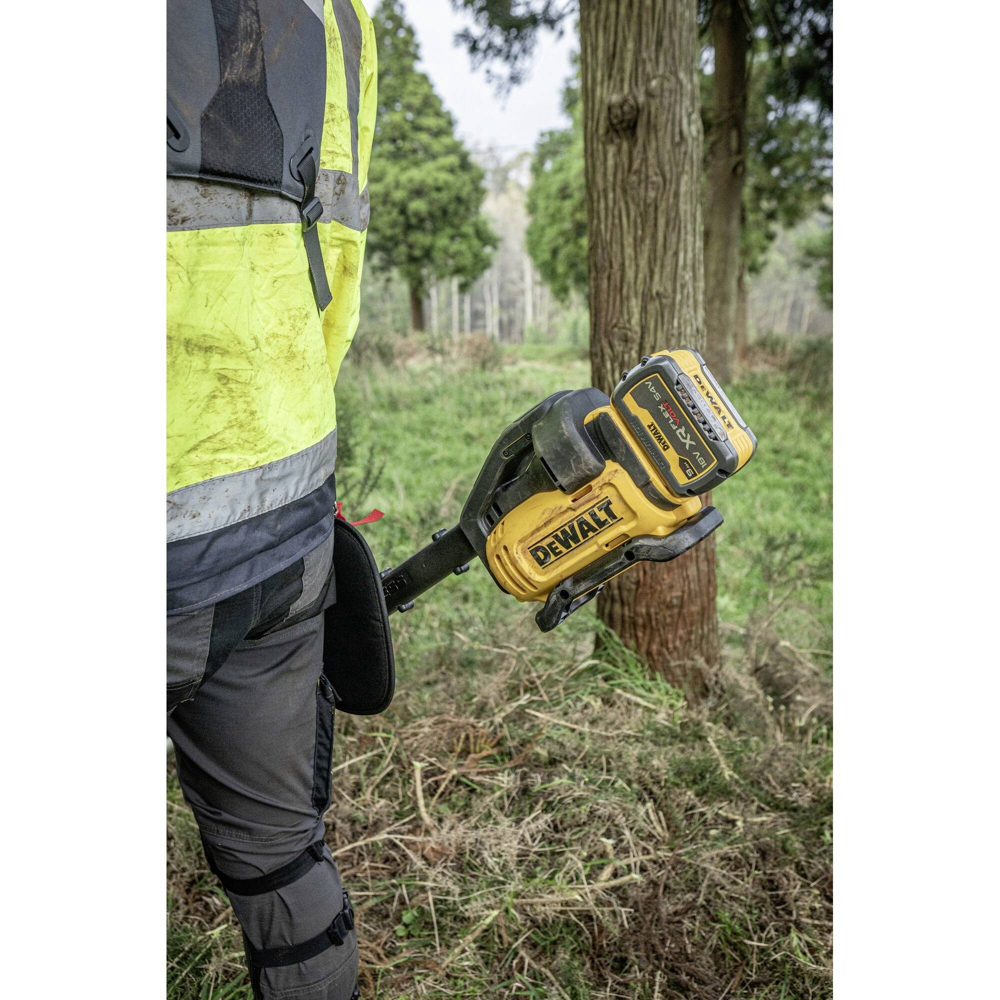 A person wearing a yellow jacket is holding a yellow, battery-powered garden tool in a wooded area.