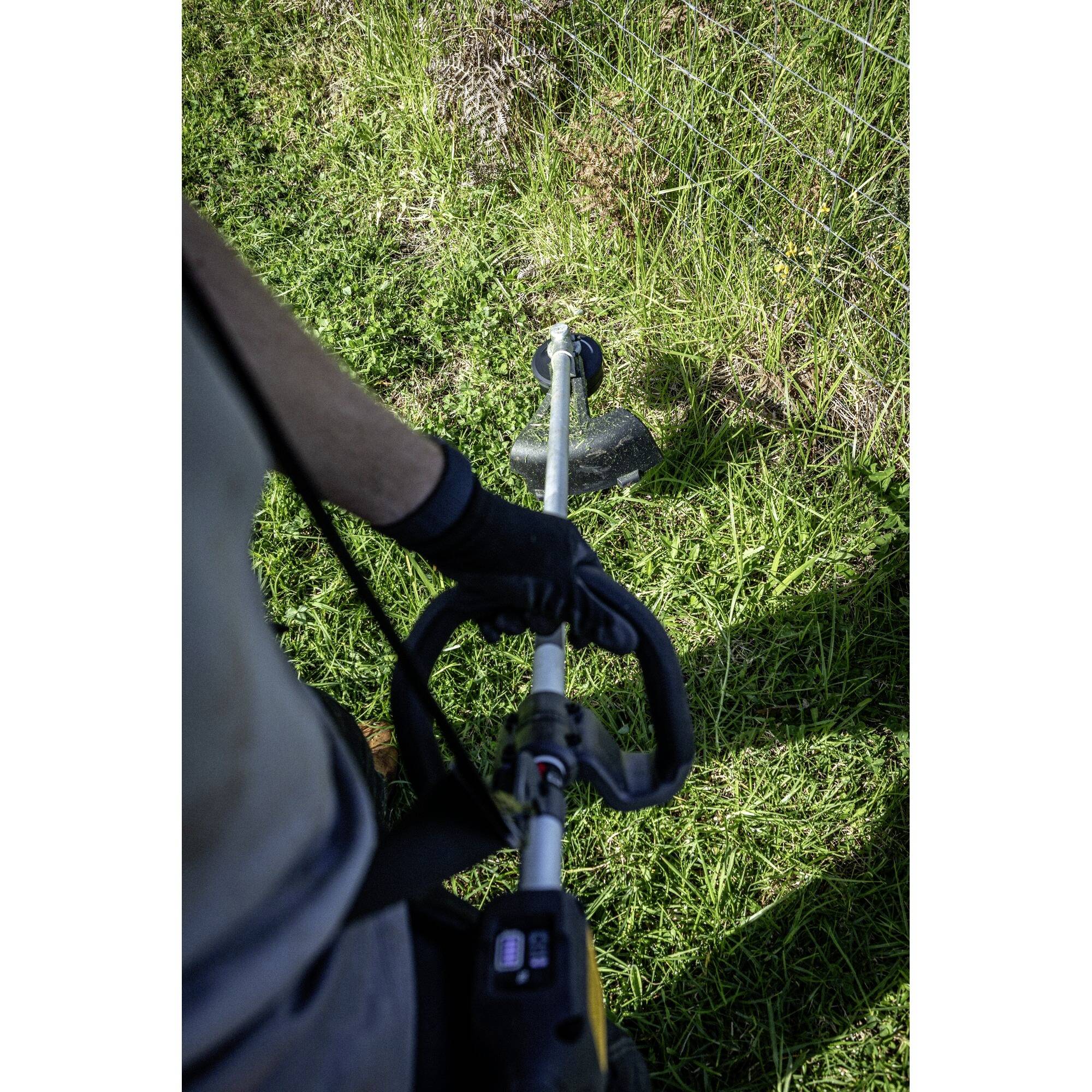A person is using a strimmer to trim grass along a fence. The scene depicts the outdoor work process.