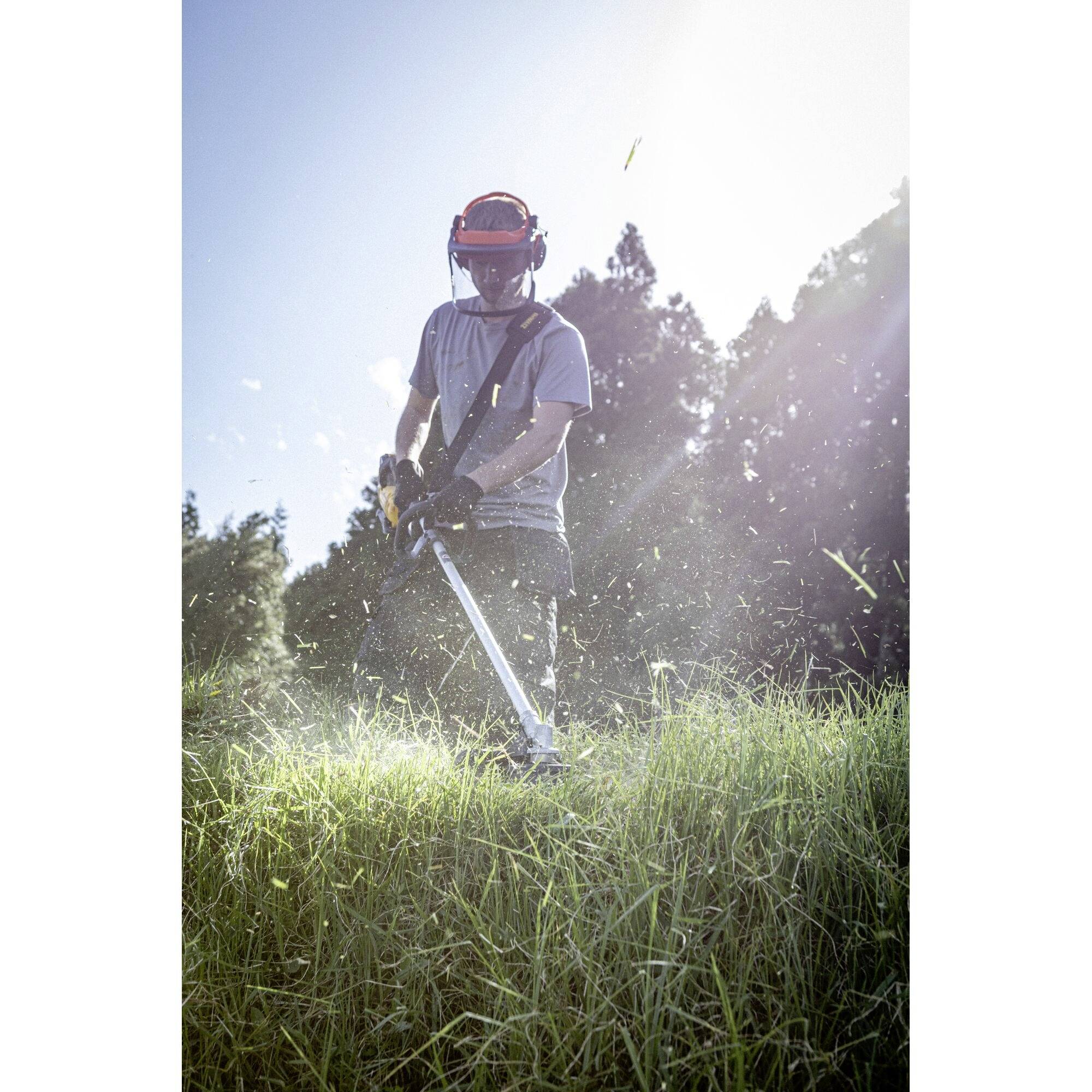 A person is mowing tall grass with a strimmer, wearing protective clothing including a helmet and ear defenders, against a wooded background.