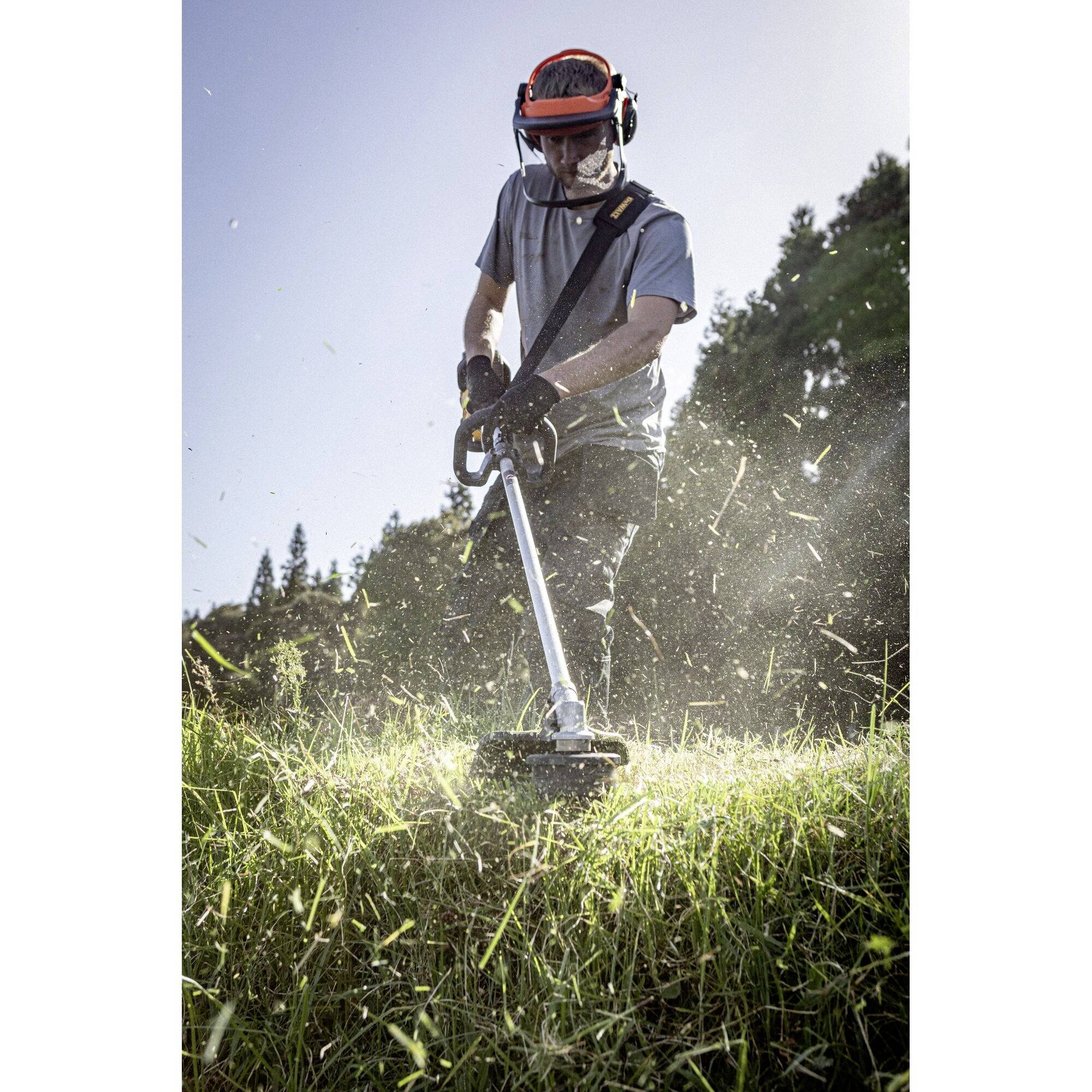 A person is mowing grass with a strimmer outdoors, wearing protective clothing. Sunlight streams through the trees.